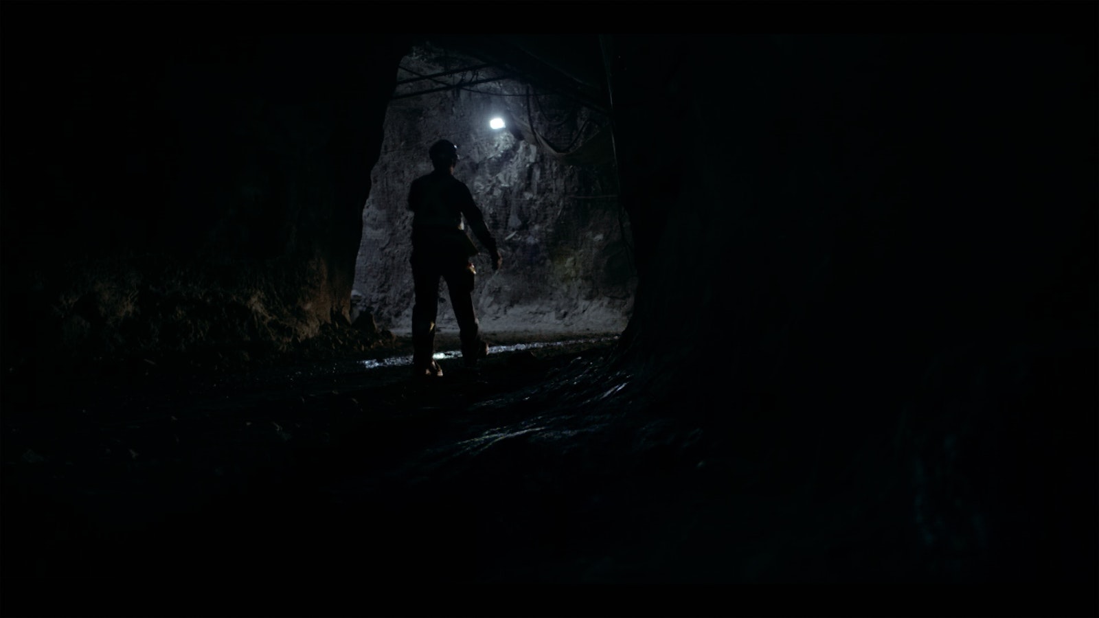 A mineworker stands in an underground tunnel surrounded by grey rock. A single white light shines in the tunnel.