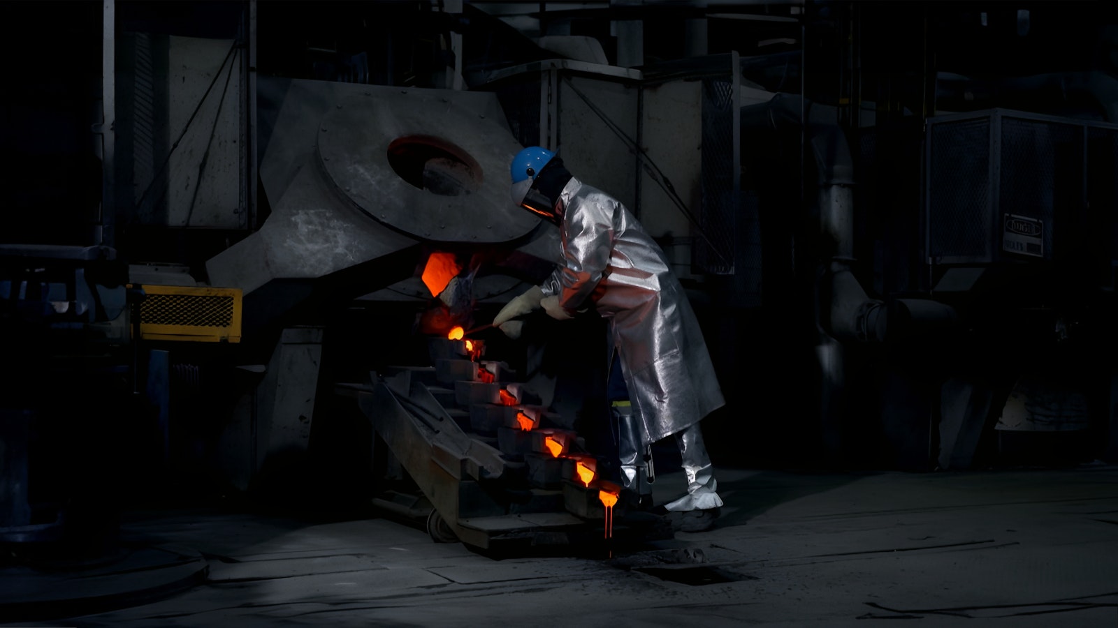 A miner, wearing silver, heat-reflective gear, arranges bar-shaped moulds at the mouth of a gold-smelting furnace.