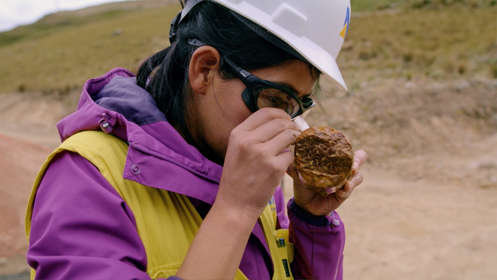Denisse Quispe inspects a large rock, a brown Earth core, with a magnifying glass in her right hand.