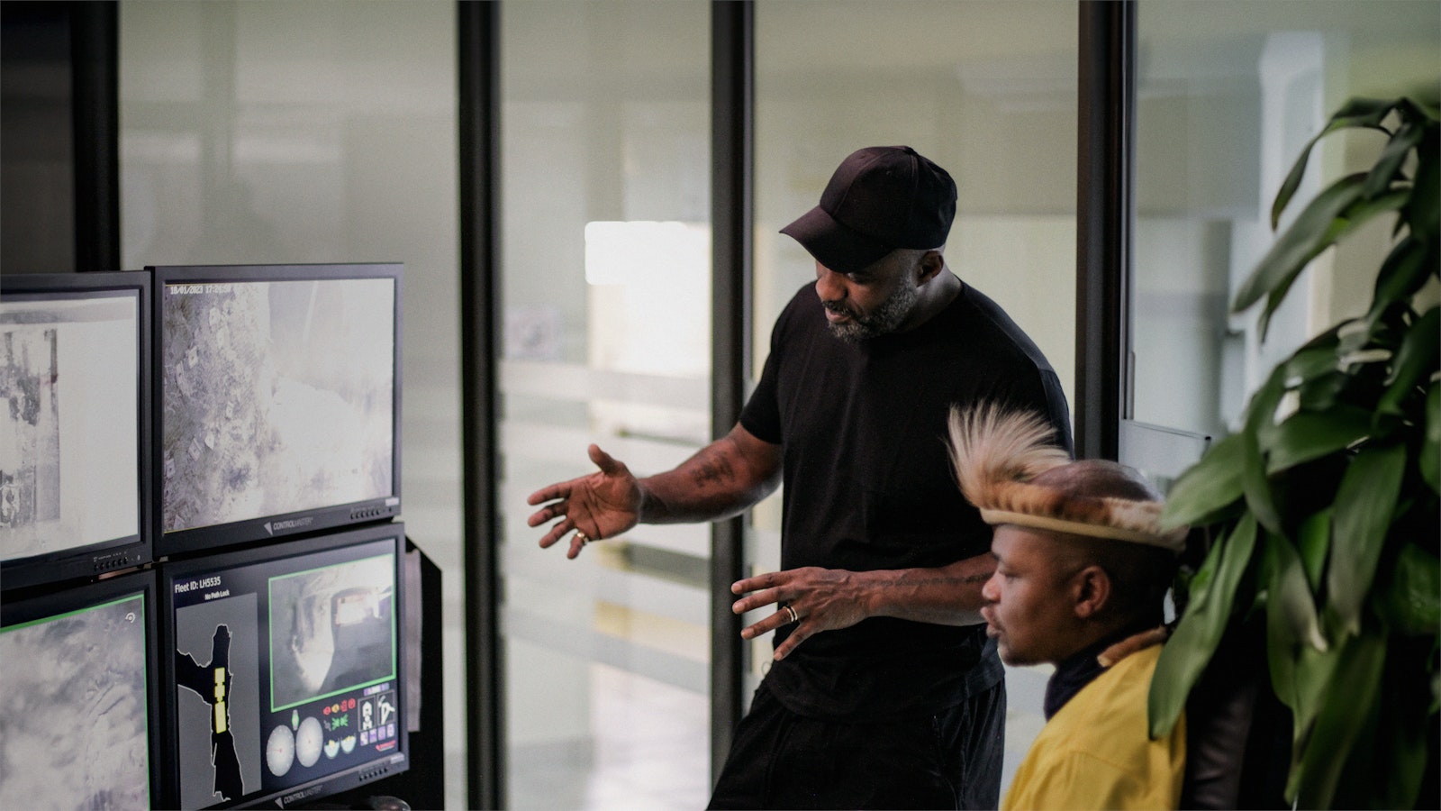 A mineworker sits and shows a standing Idris Elba the console where he controls mine machinery remotely.