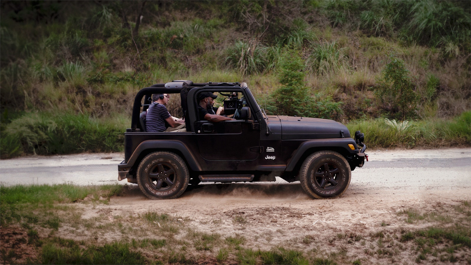 Idris Elba drives a back 4x4 car on a road through green of the South African farmland with a cameraman in the back.