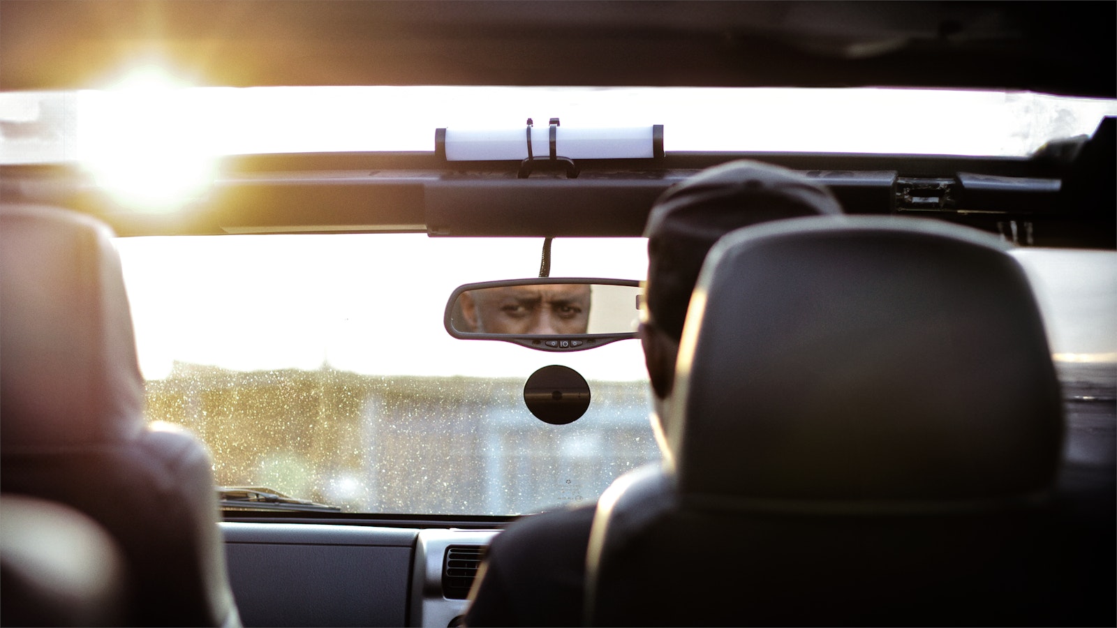 A shot of Idris Elba from a car’s back seat. The sun shines into the windshield, Idris’ eyes reflect in the rearview mirror.