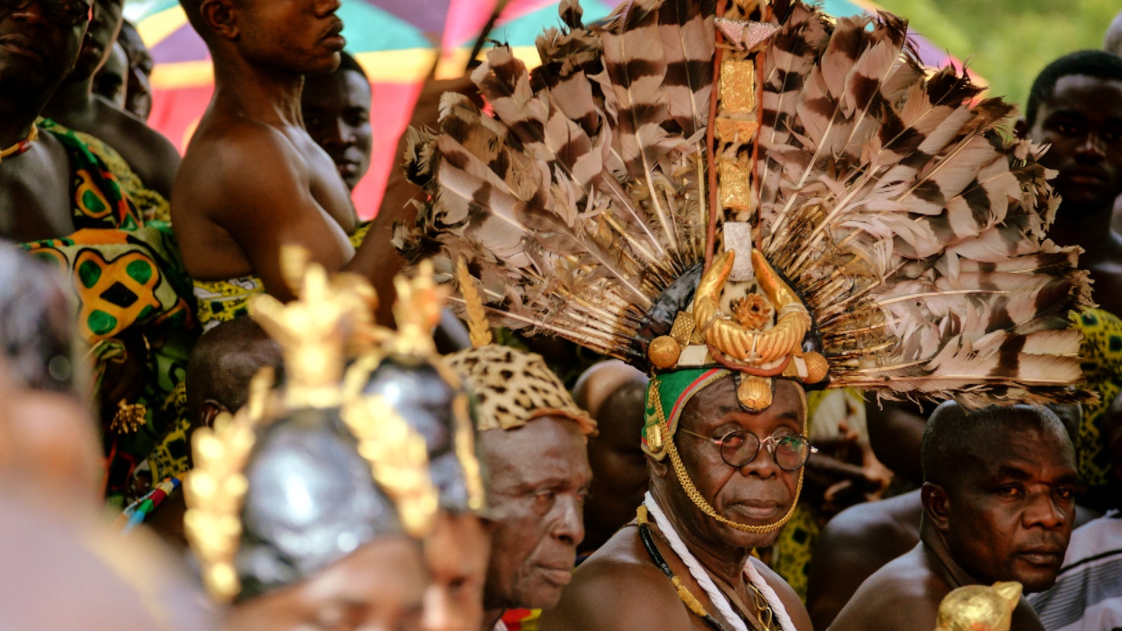 The chiefs await meeting the king. The chief in focus wears a large fan-shaped head adornment covered with gold details.