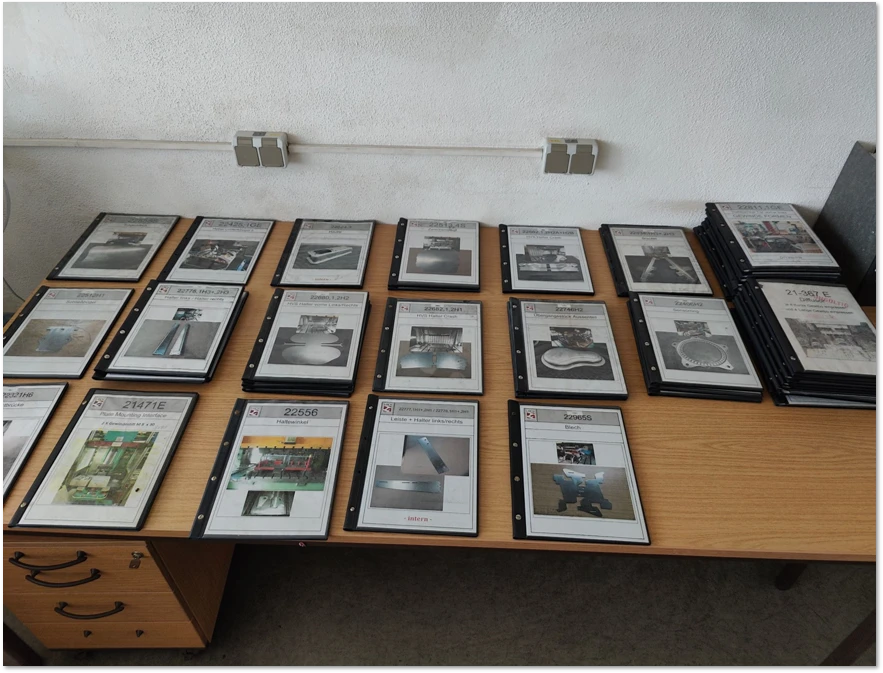 Zahlreiche Aktenordner mit eingelegten Dokumenten und Produktfotos liegen aufgereiht auf einem Bürotisch.