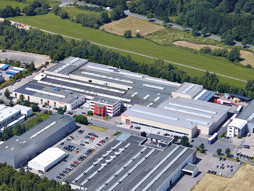 Aerial view of a large industrial site with several production and warehouse buildings, parking lots, and surrounding fields. Bird's-eye photo of a factory campus showing elongated hall roofs, a multi-story office building in the middle, and numerous vehicles in the surrounding parking areas. Overview shot of a manufacturing site illustrating the spatial layout of shopfloor halls, logistics areas, and adjacent infrastructure.