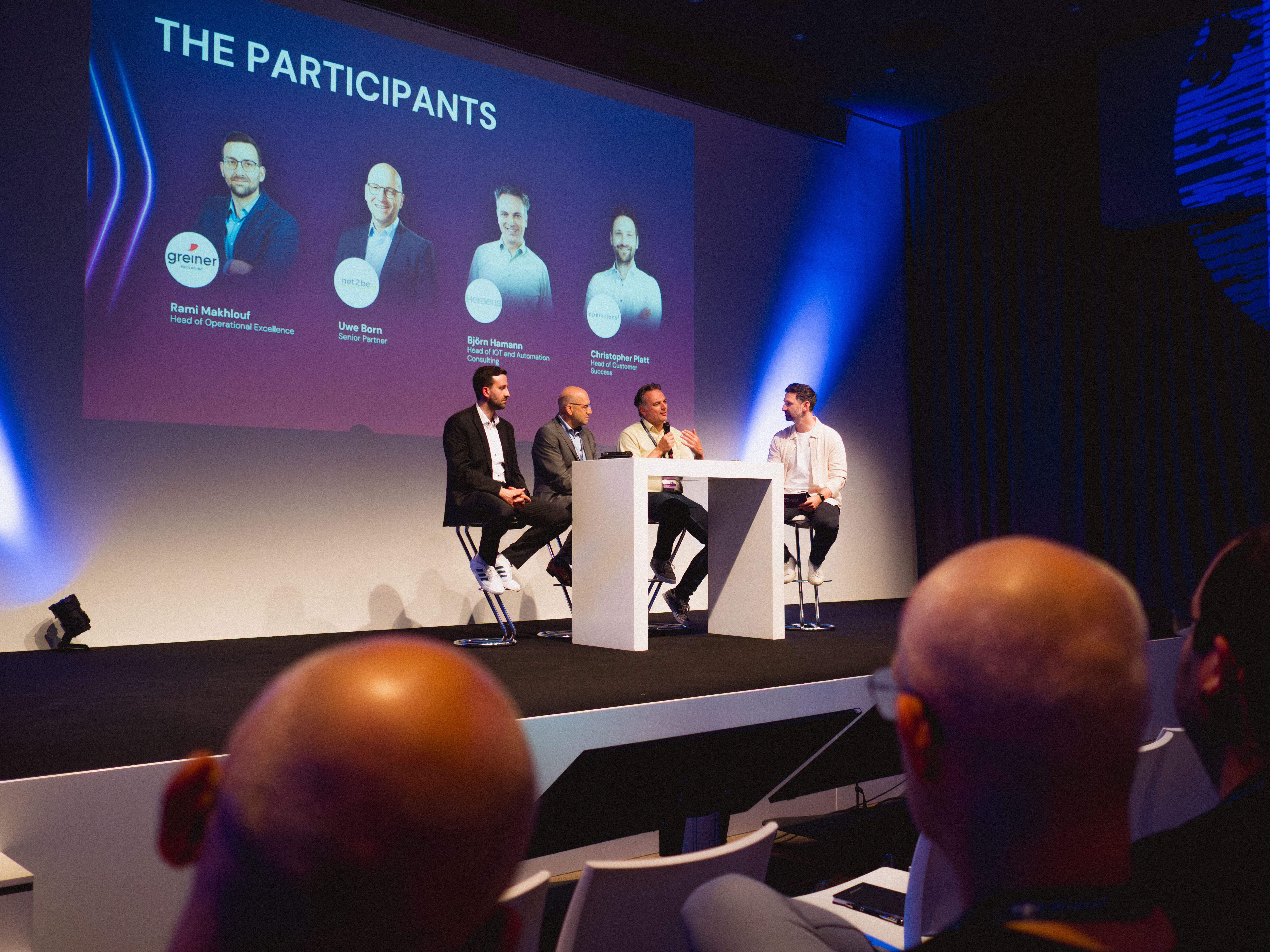 Podiumsdiskussion mit den vier Teilnehmern, während einer mit Mikrofon spricht und die Leinwand ihre Namen und Funktionen wie Head of Operational Excellence und Head of Customer Success zeigt.