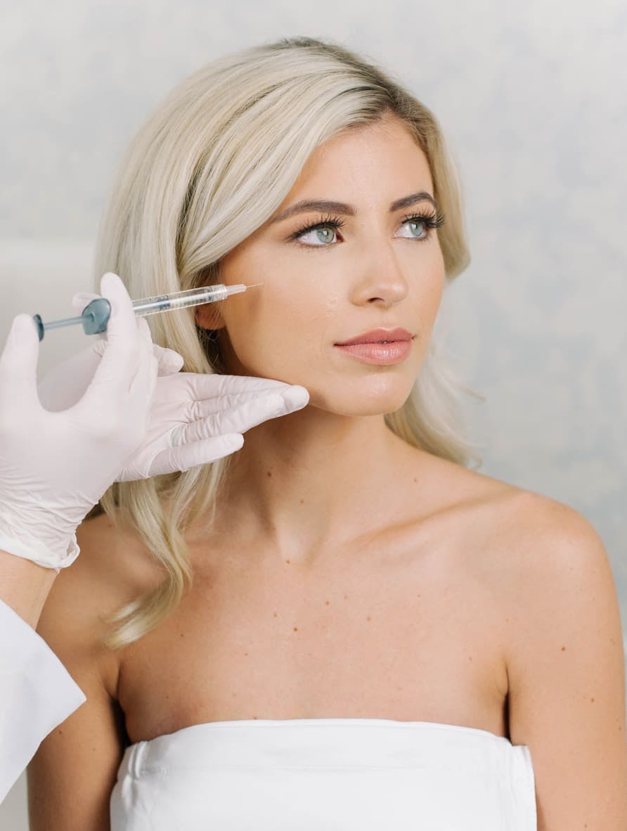 Blonde woman looking to the right as gloved hands hold a needle to her face