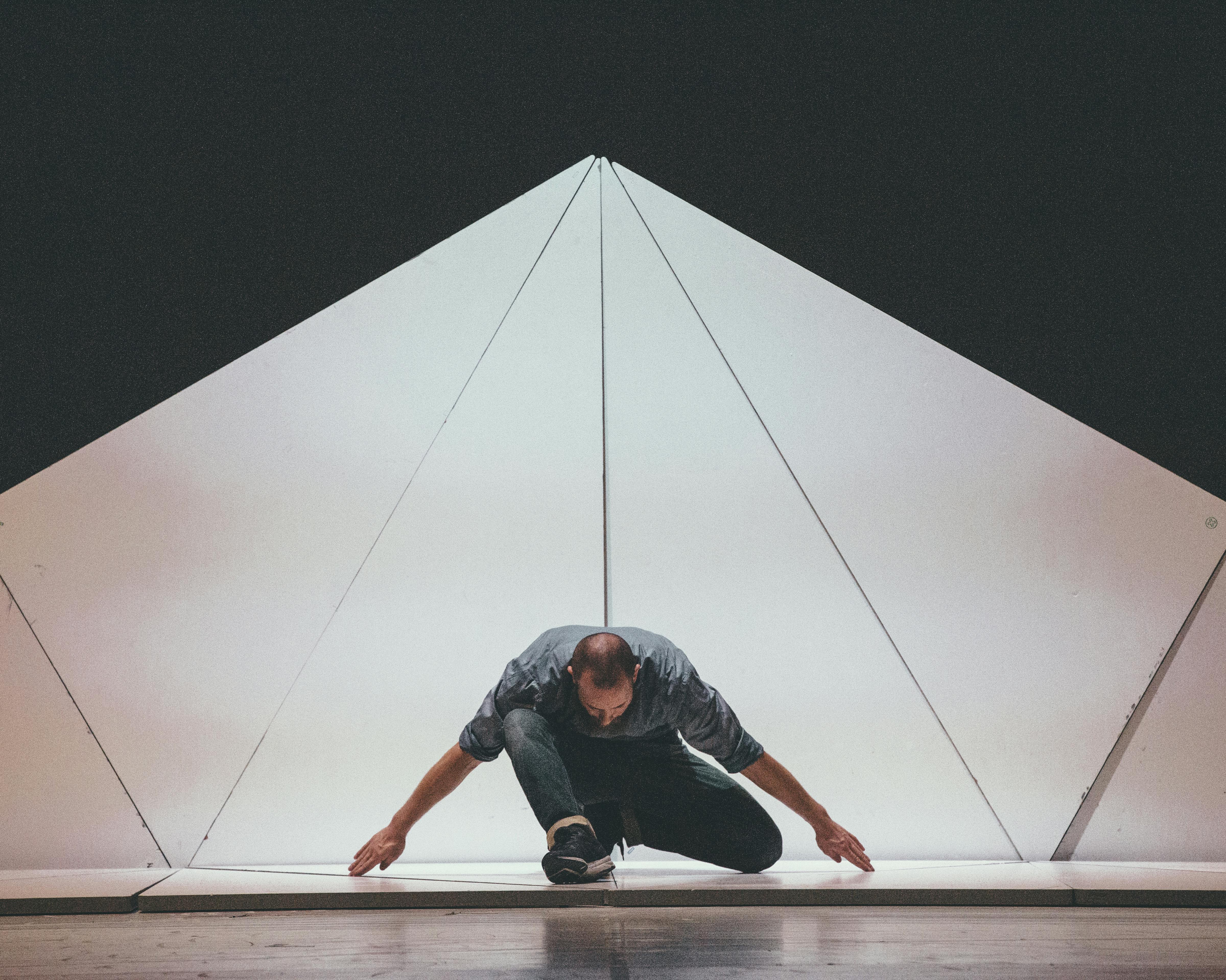 At the centre of the stage, a dancer crouched on the ground. Behind, a white pyramid-shaped canvas on a black background.