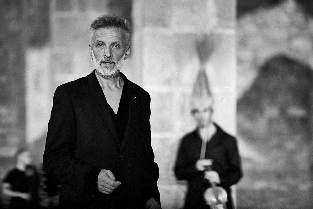 Pep Ramis is in the foreground. Half-length, standing, he is facing the camera, his right arm on his stomach and his left arm along his side. Behind a girl is holding a violin vertically along her stomach.