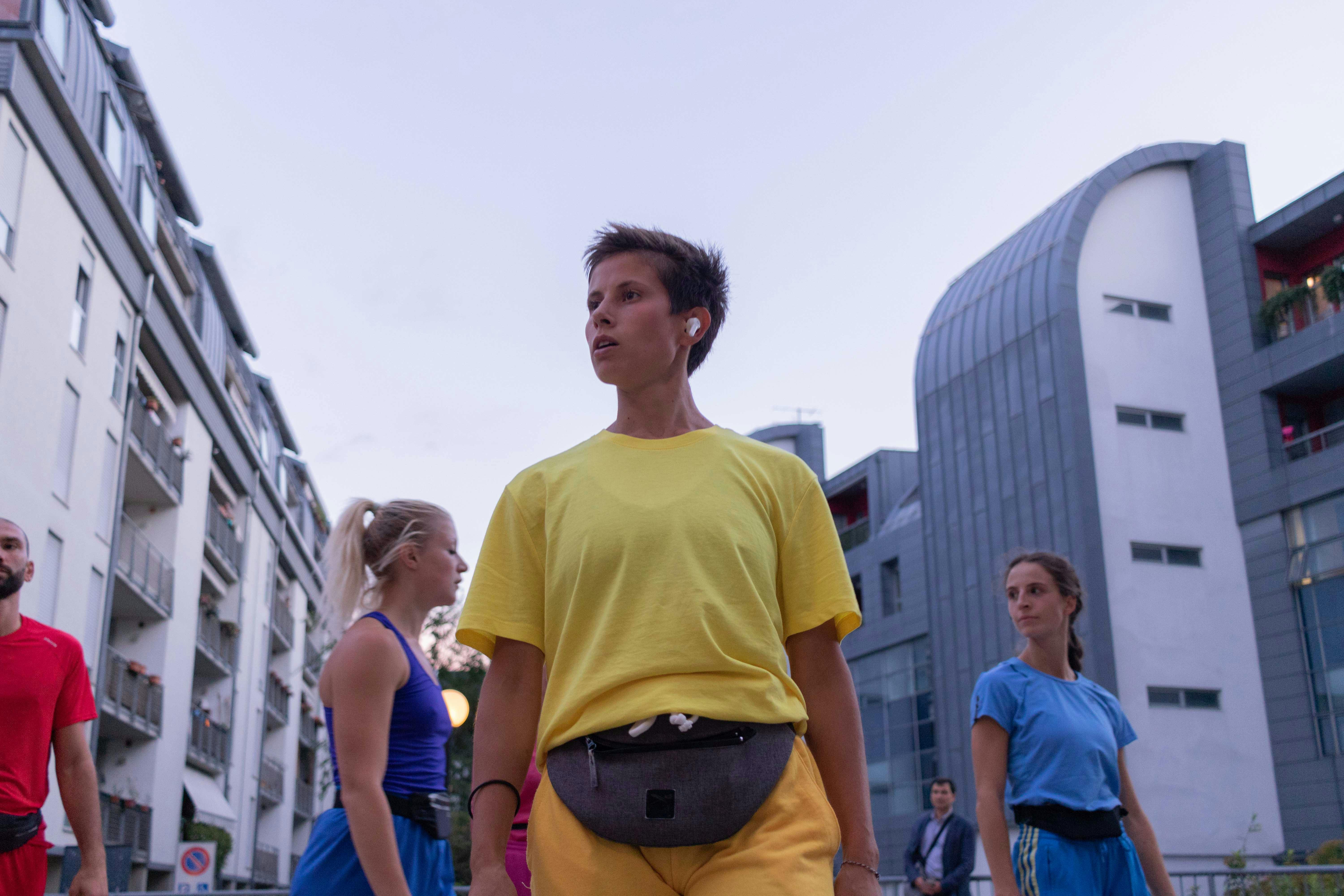 During a rehearsal in a square, a girl stands in front of the camera. She keeps her arms at her sides and her gaze pointed towards a point outside the lens.