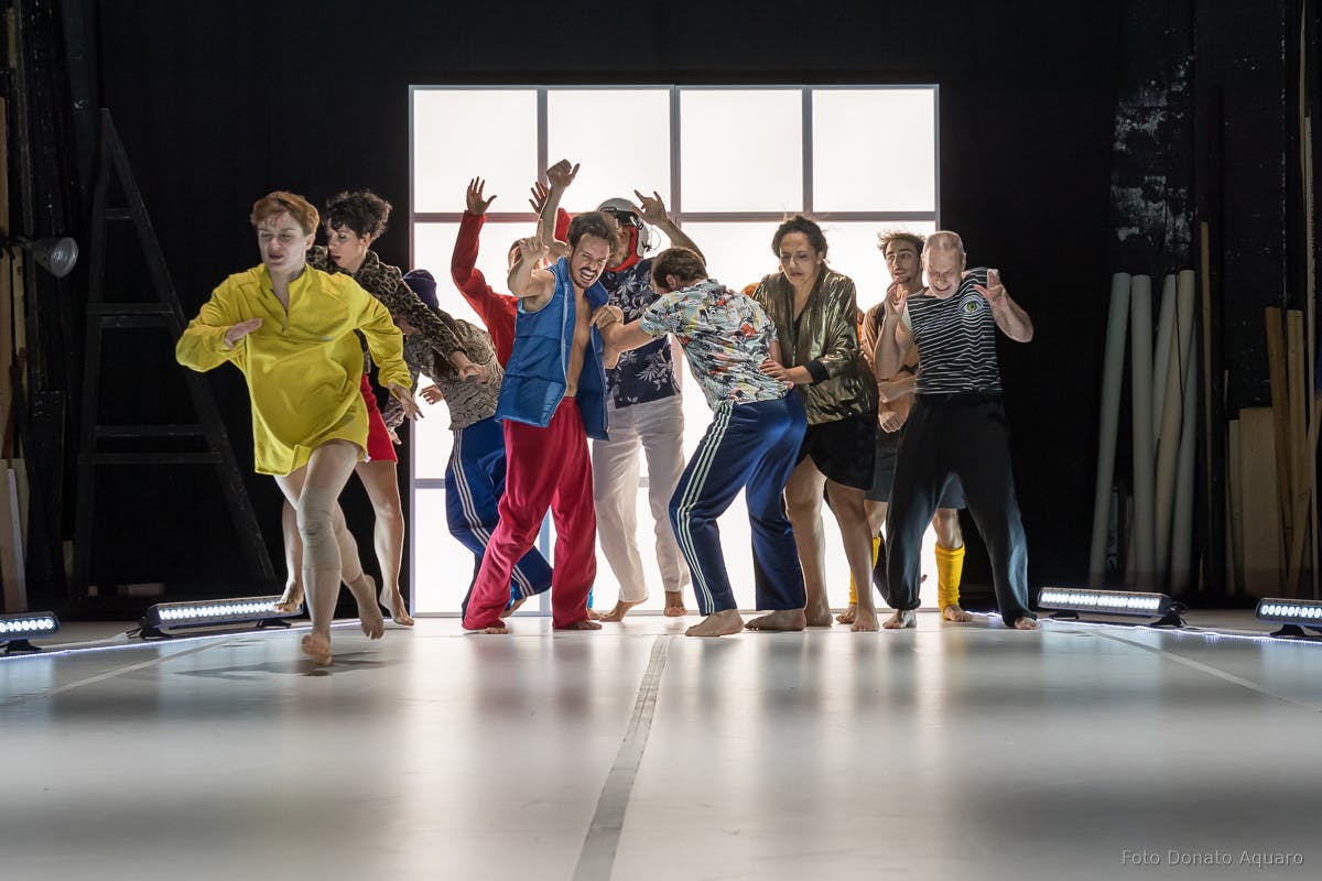 The group of dancers is at the back of the room and ready for rehearsals, while a leading dancer leads.