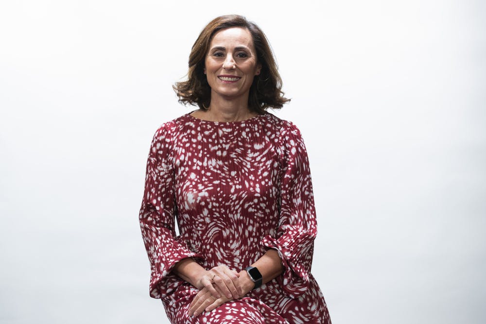 Photo of Laura Canali seated on a stool. Her face is turned towards the camera in a smile, while her joined arms are gathered in her lap.