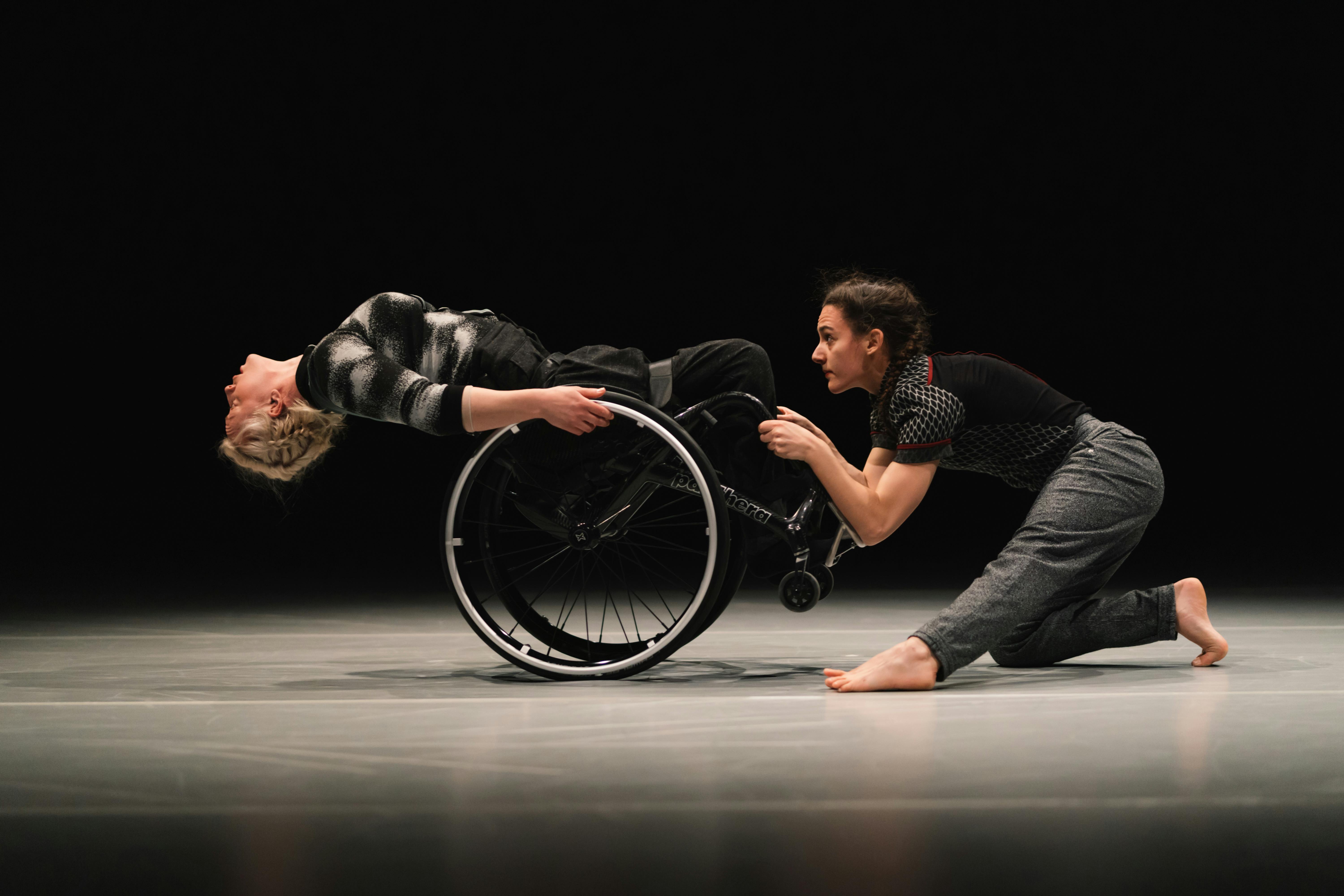 Two dancers, one of them in a wheelchair are in profile facing each other. The dancer in the wheelchair is falling backwards, while the other grabs her legs.