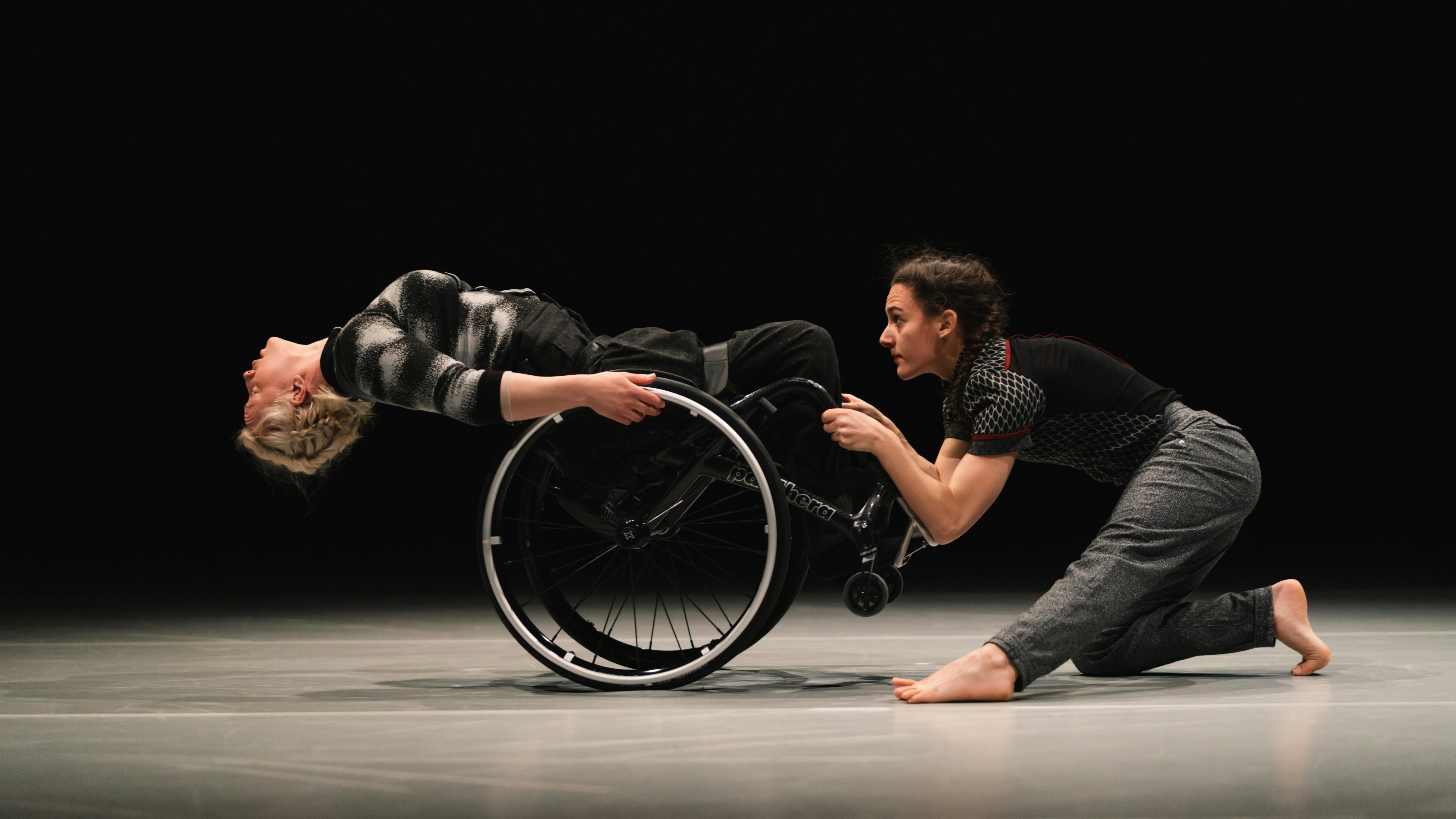 Two dancers, one of them in a wheelchair are in profile facing each other. The dancer in the wheelchair is falling backwards, while the other grabs her legs.