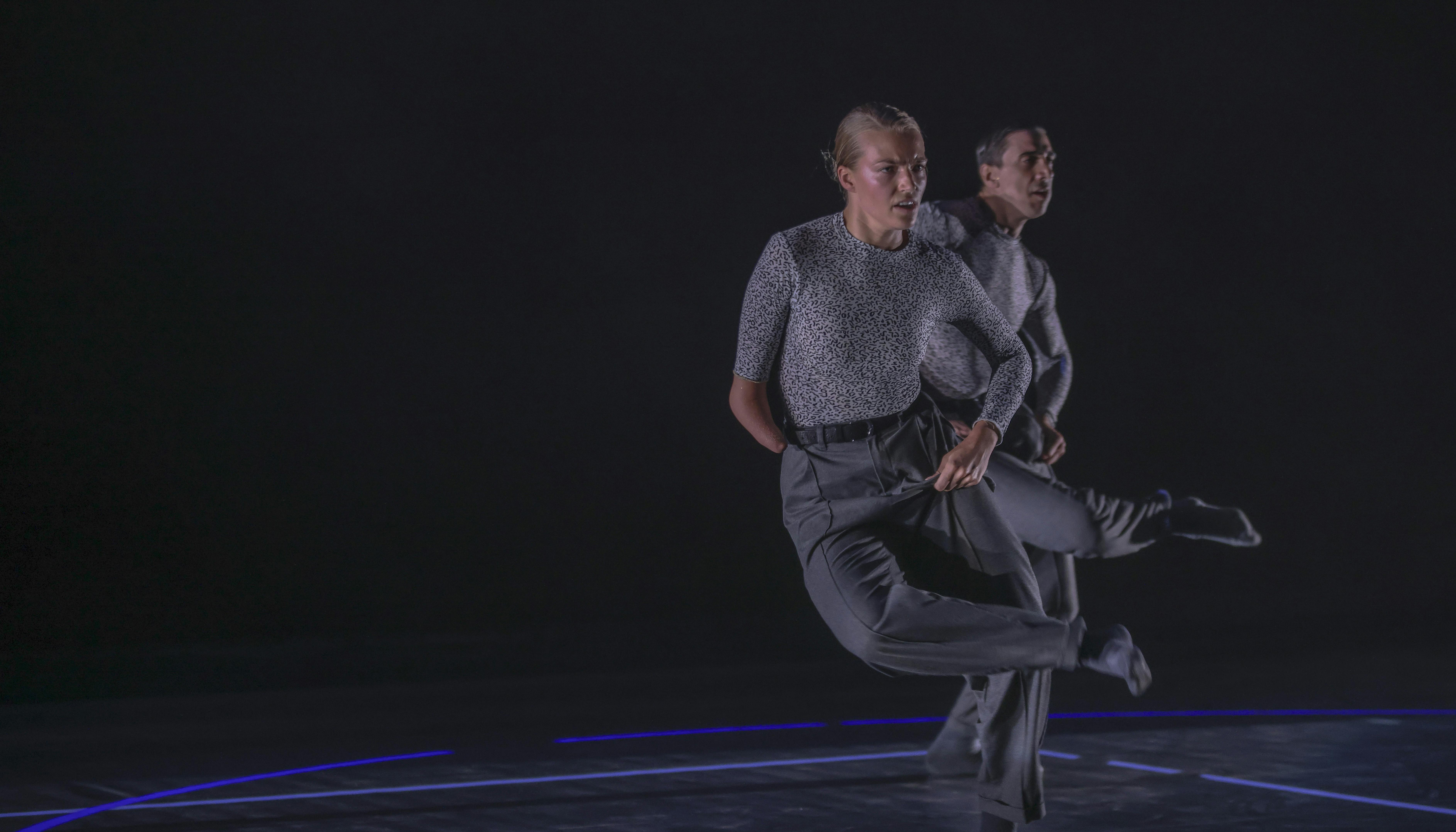 Sul palco un danzatore piega la gamba destra mettendo il piede sul ginocchio opposto. Davanti a lui, una danzatrice con disabilità fa lo stesso movimento.