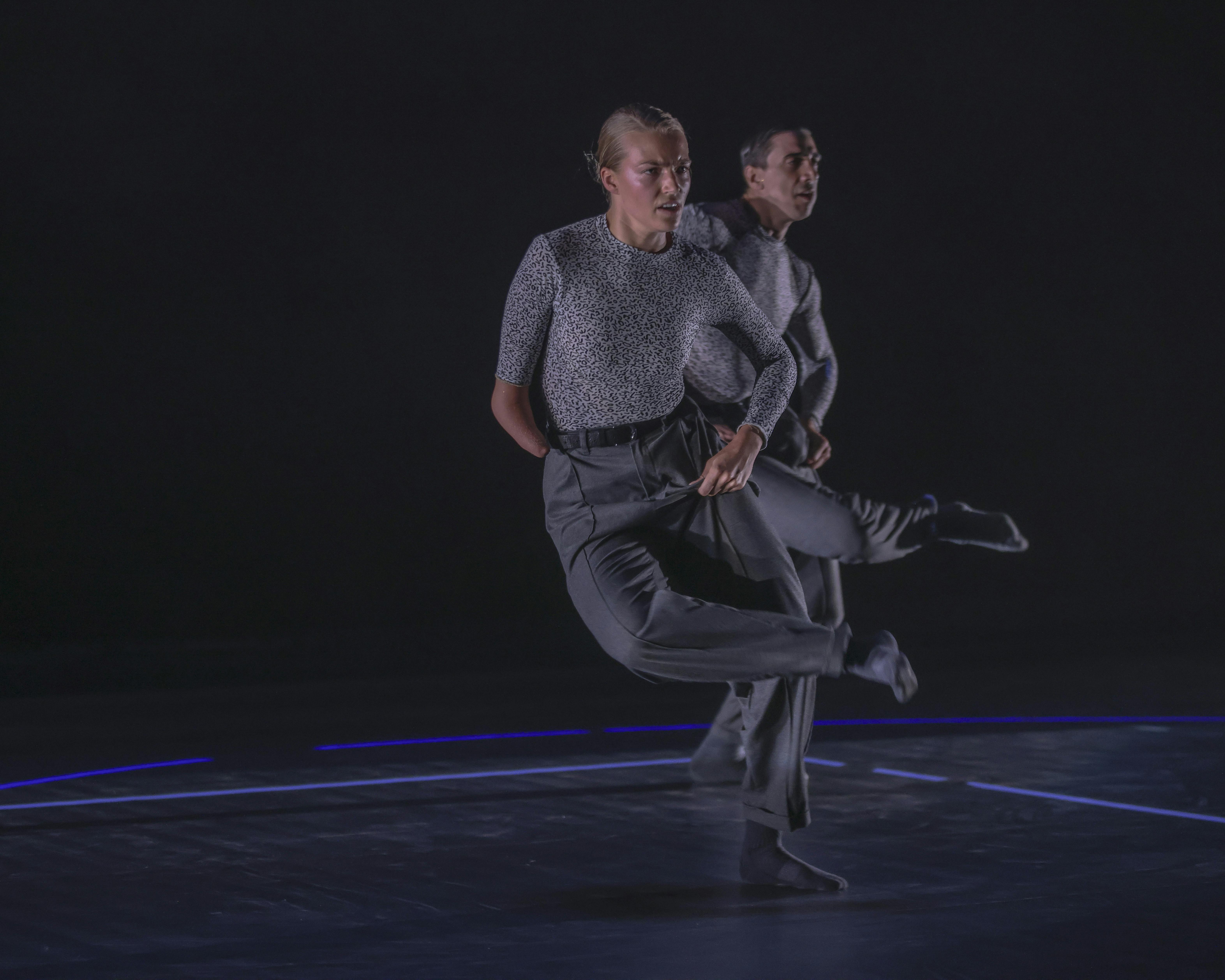 On stage, a dancer bends his right leg by placing his foot on the opposite knee. In front of him, a dancer with a disability does the same movement.
