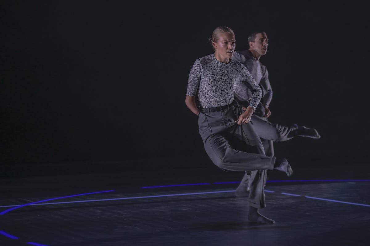 Sul palco un danzatore piega la gamba destra mettendo il piede sul ginocchio opposto. Davanti a lui, una danzatrice con disabilità fa lo stesso movimento.