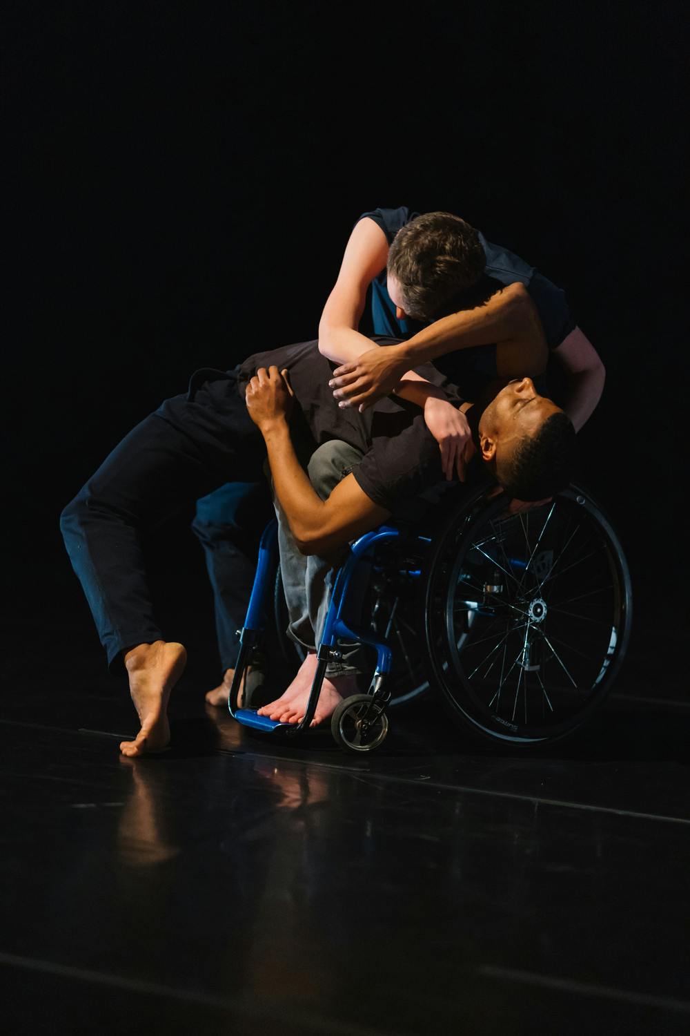 In the center of the scene, a standing dancer arches his back until he falls onto the legs of a dancer in a wheelchair, who then rests his head on his chest and wraps his arms around him.