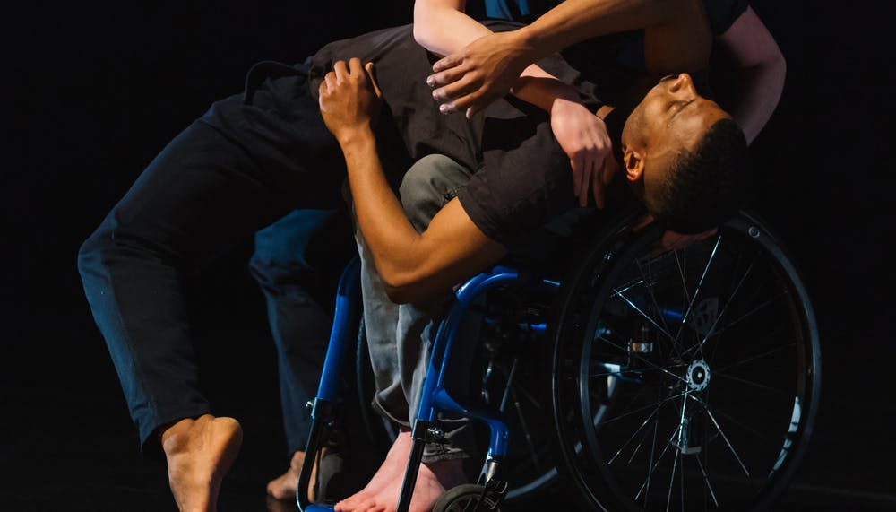 In the center of the scene, a standing dancer arches his back until he falls onto the legs of a dancer in a wheelchair, who then rests his head on his chest and wraps his arms around him.