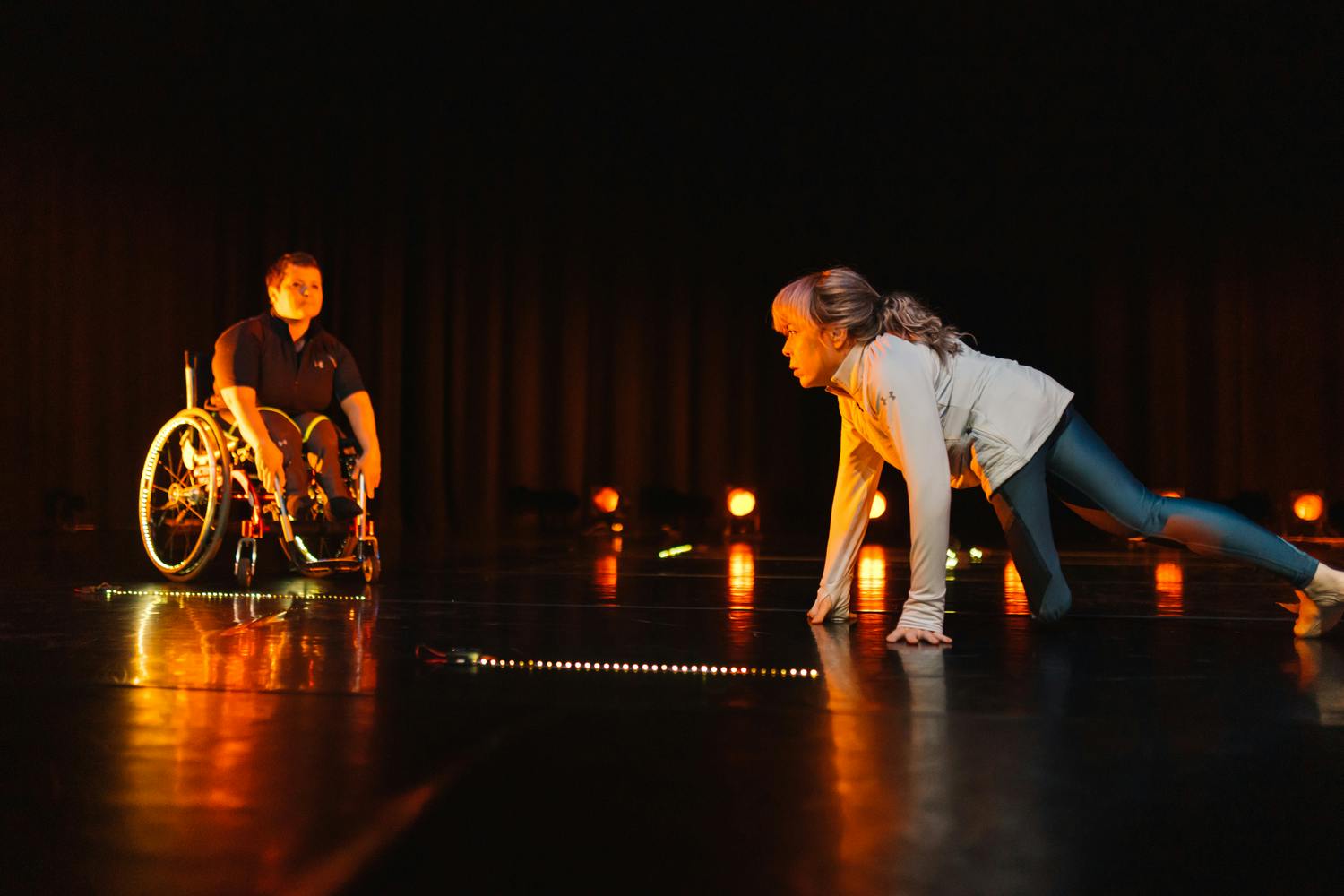 On stage are two female performers with disabilities, one in a wheelchair and the other on crutches. On the right, having abandoned her crutches, the dancer moves across the floor toward the dancer in the wheelchair.