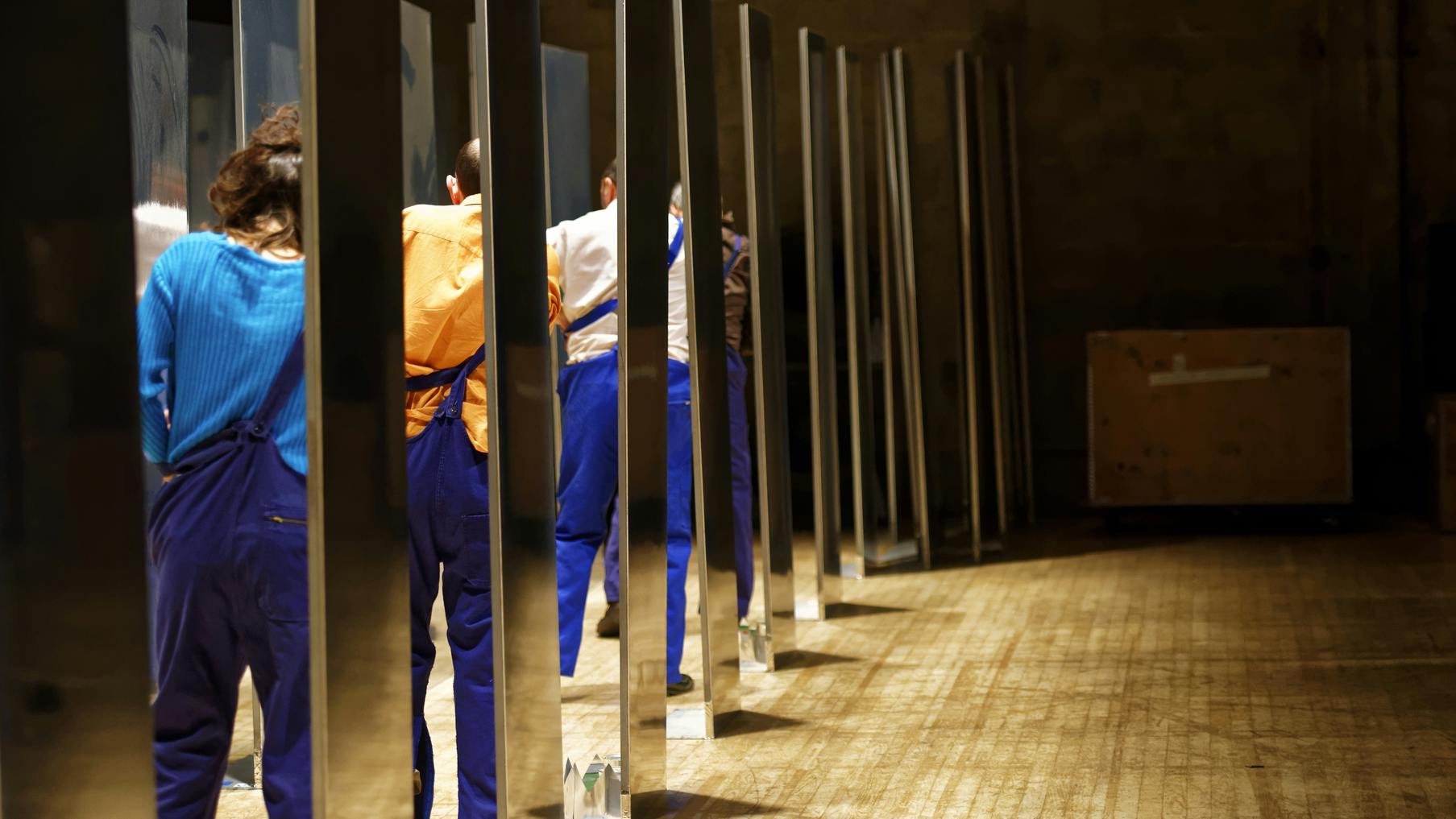Sul palcoscenico sono stati adibiti dei camerini, visti lateralmente, dove all'interno ci sono i danzatori in piedi e di spalle nell'intento di tirare su la bretella sinistra dei pantaloni.
