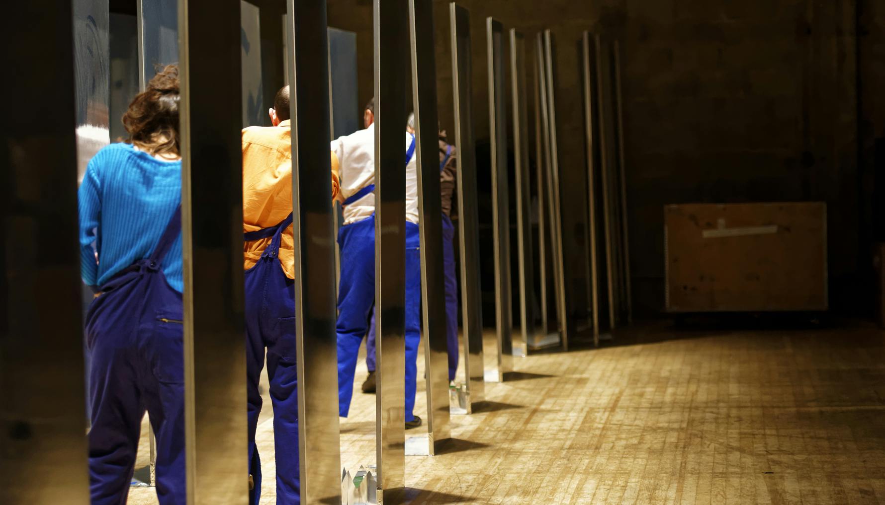On the stage there are dressing rooms, seen from the side, where the dancers stand with their backs to the stage and try to pull up the left suspender of their trousers.