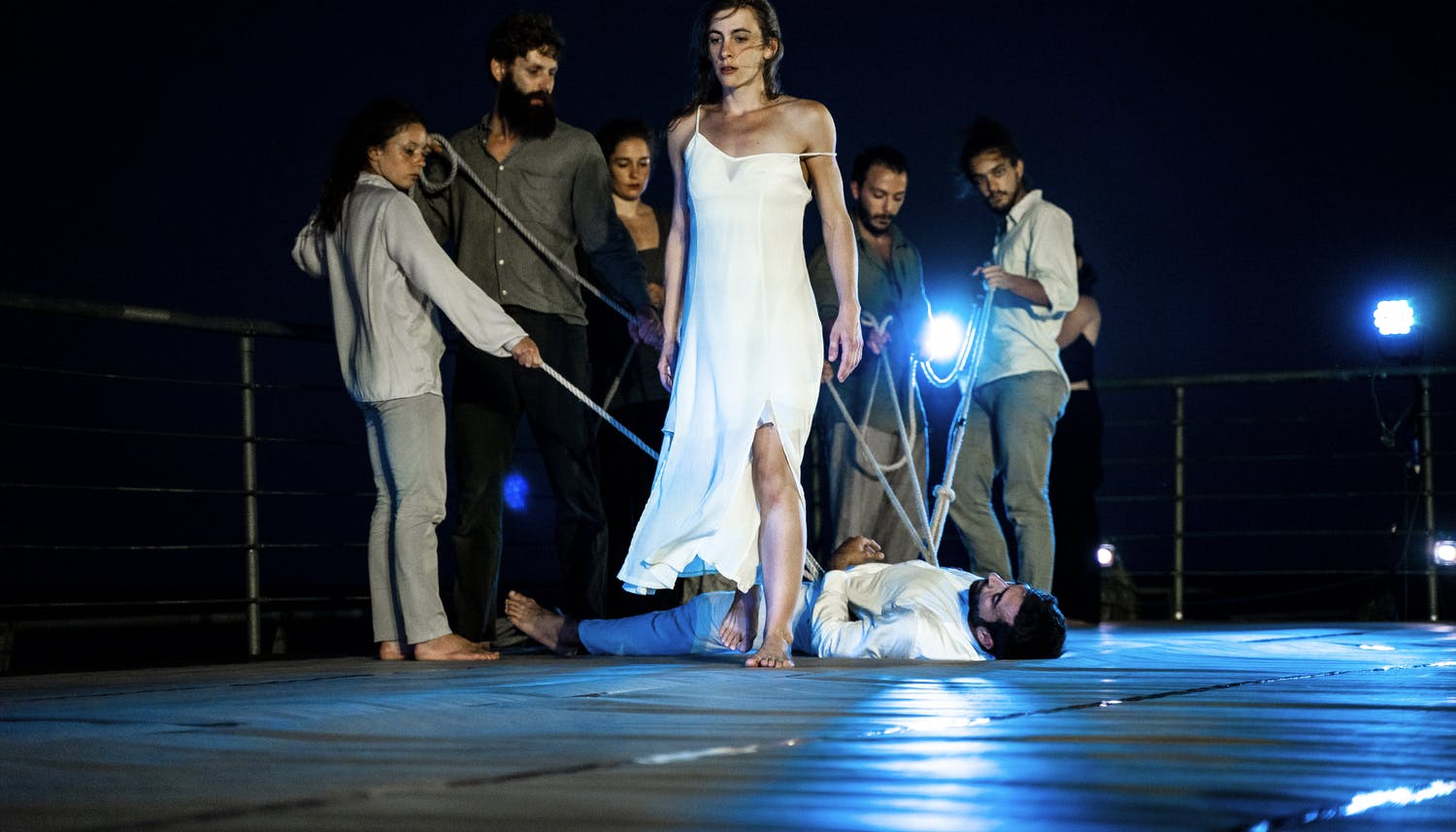 Una danzatrice da le spalle a un gruppo di danzatori che tengono legato a terra un uomo steso.