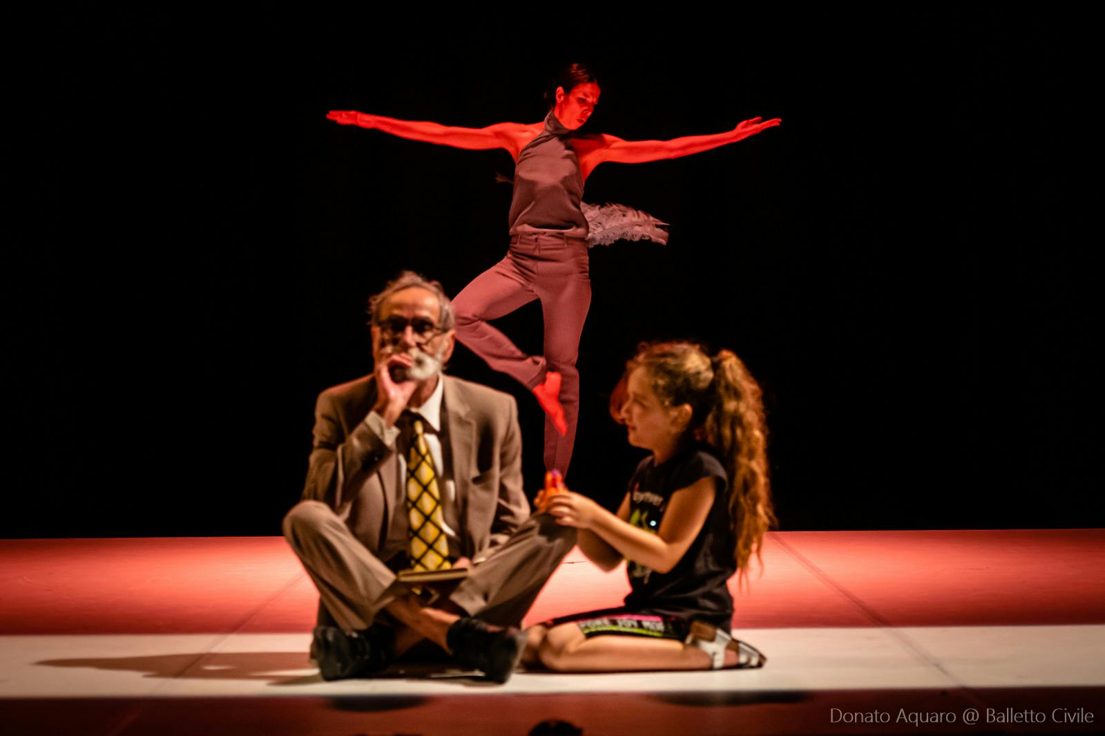 In prima fila il padre di Michela Lucenti seduto a gambe incrociate con una ragazzina che siede a fianco sulle ginocchia ed è rivolta verso di lui. Dietro una ballerina classica sta sulle punte con le braccia allargate ad altezza spalle.