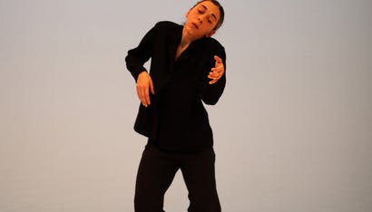 María Muñoz in piedi al centro del palcoscenico danza con le gambe incrociate, piede destro in avanti, braccia alzate ad altezza del petto.