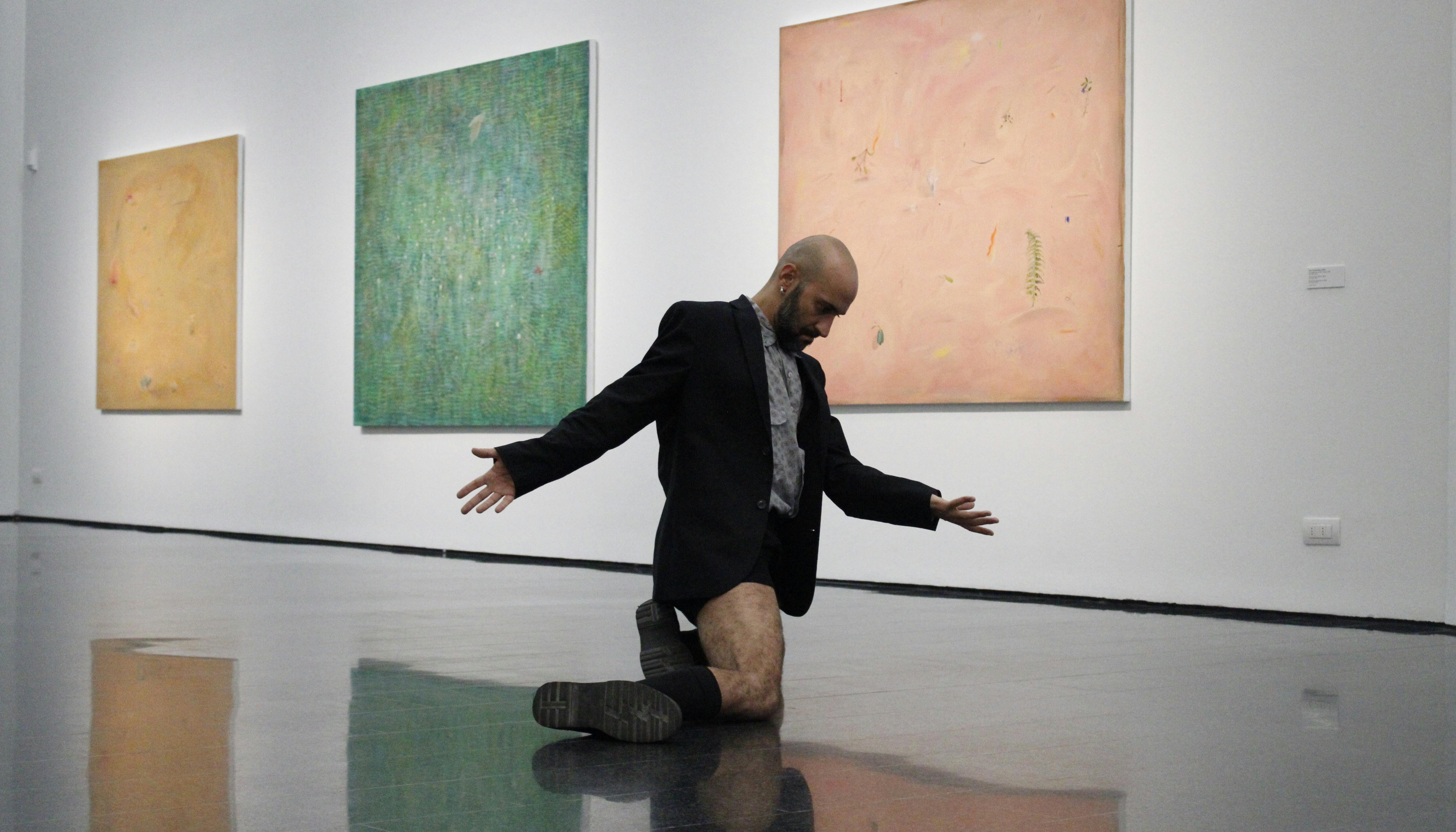 Daniele Ninarello è inginocchiato, le braccia leggermente aperte e il capo reclinato allo sterno. Dietro di lui, sulla parete, tre quadri astratti di differenti colori.