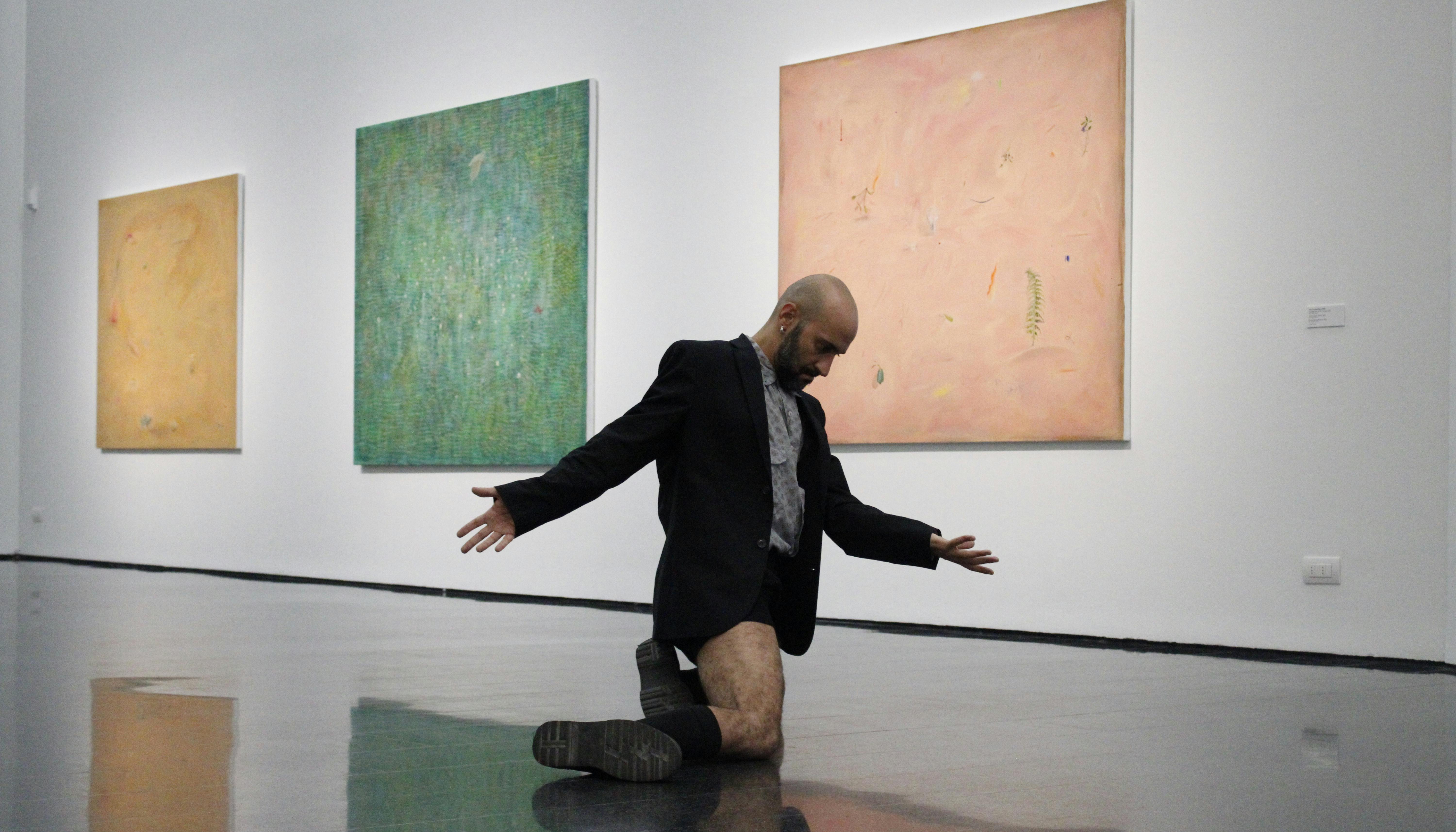 Daniele Ninarello is kneeled, his arms slightly open and his head reclined to his sternum. Behind him, on the wall, three abstract paintings of different colors.