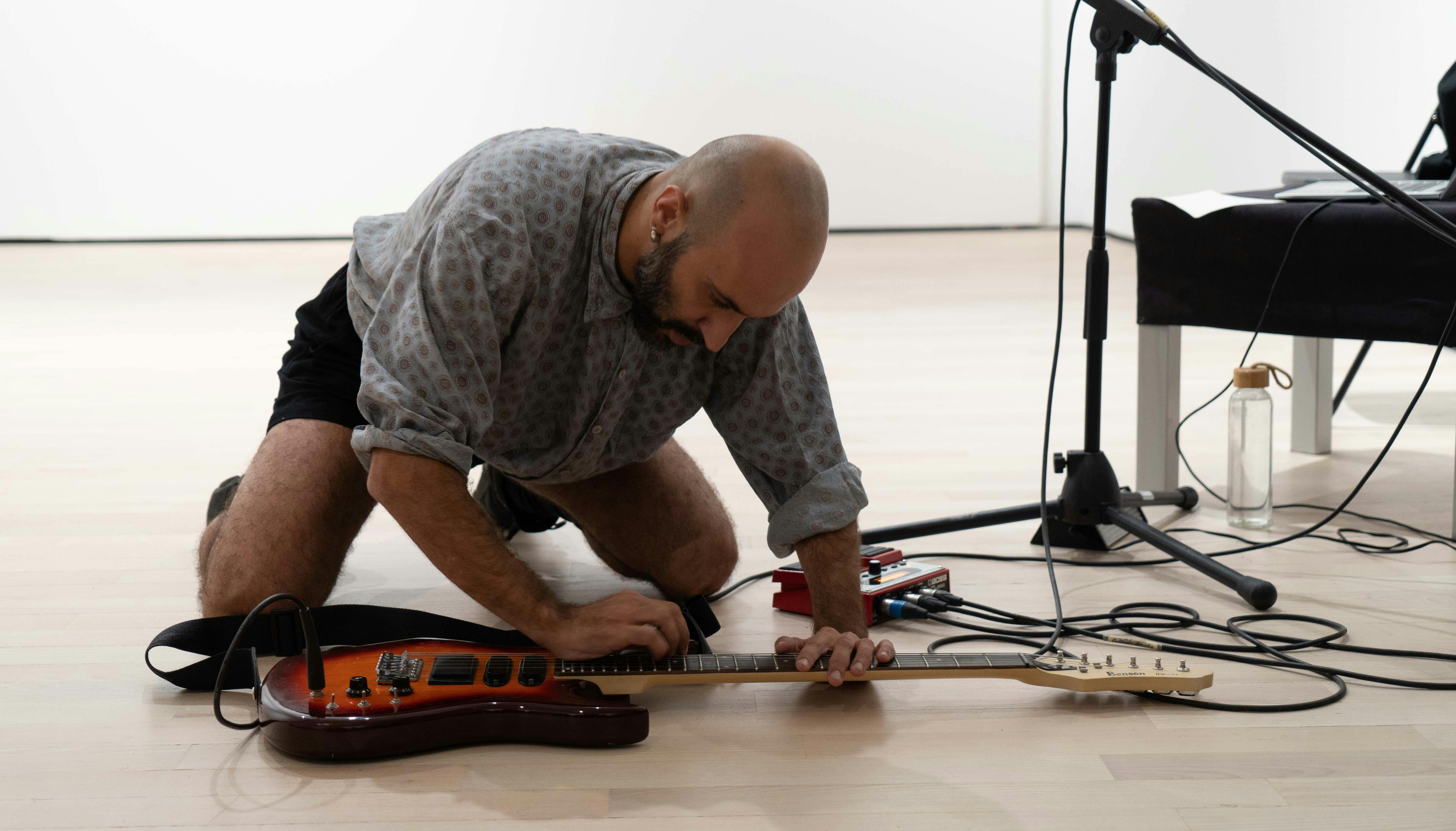 Daniele Ninarello, inginocchiato, suona una chitarra elettrica appoggiata a terra. Di fianco a lui ci sono un tavolino  nero e un asta per microfono.