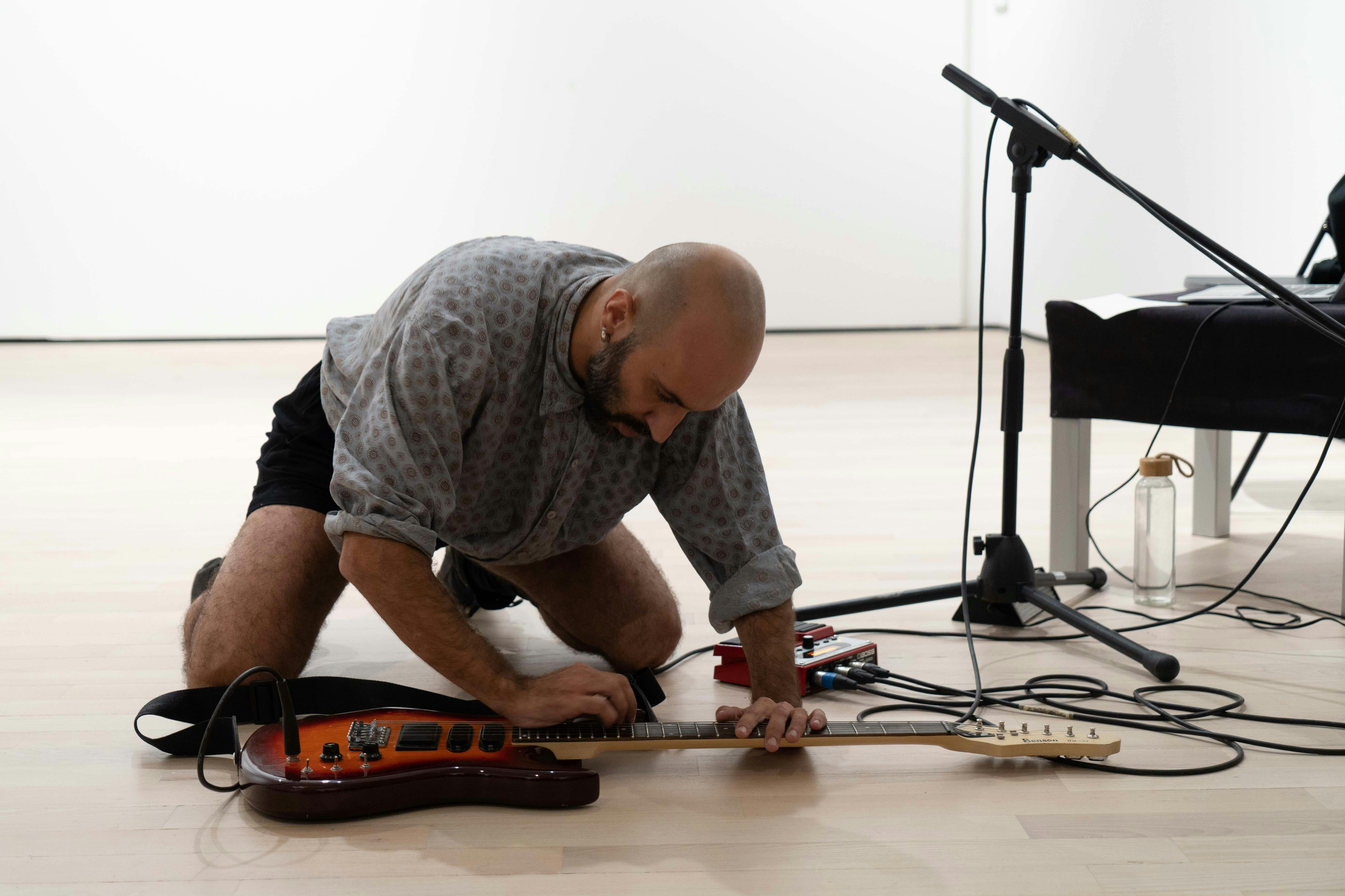 Daniele Ninarello, kneeled, plays an electric guitar laid on the ground. Next to him there are a black table and a microphone stand.