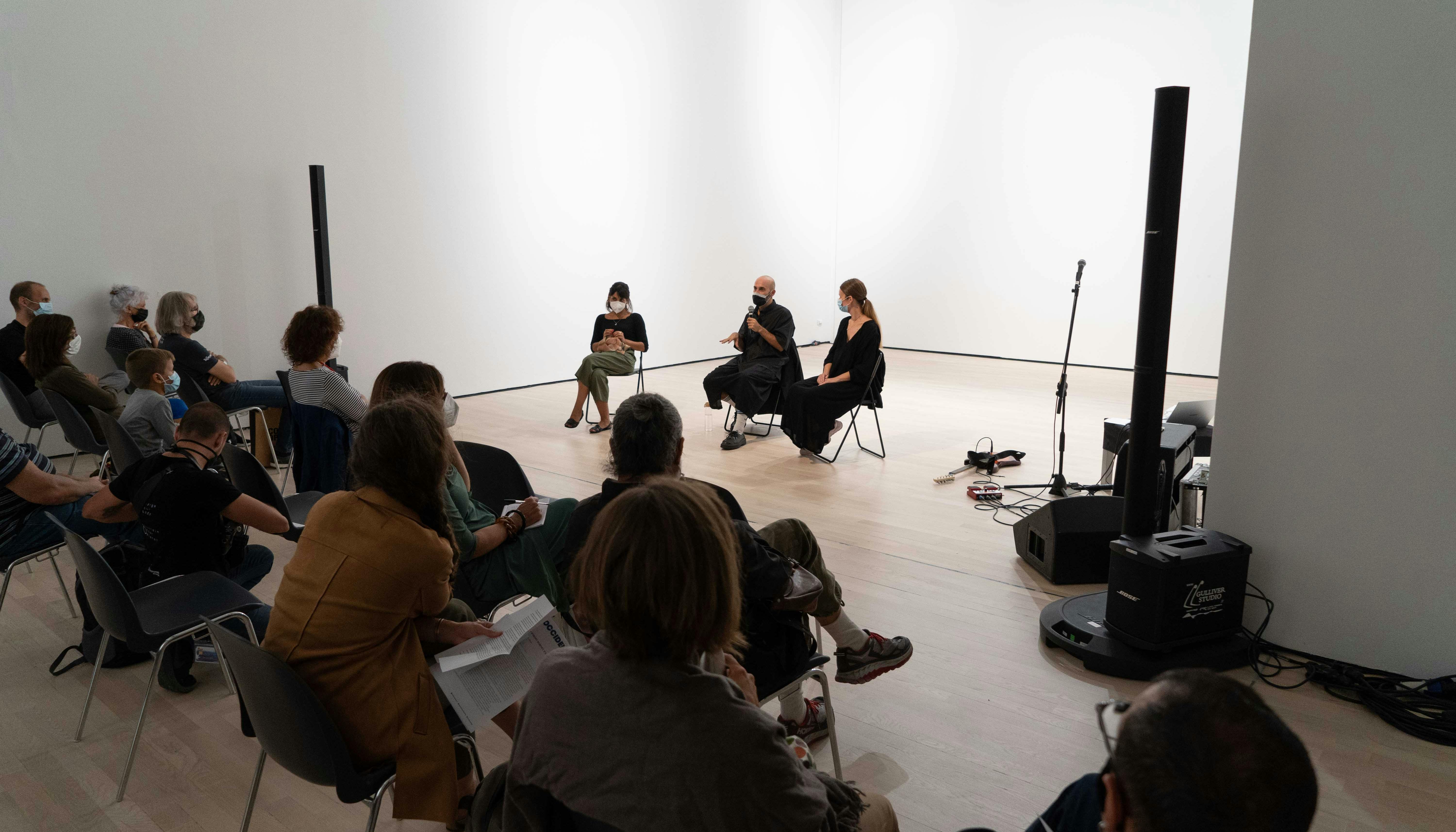 Il coreografo Daniele Ninarello, la sociologa Mariella Popolla e la drammaturga Gaia Clorilde Chernetich seduti al centro dello spazio performativo, parlano con il pubblico di fronte a loro.