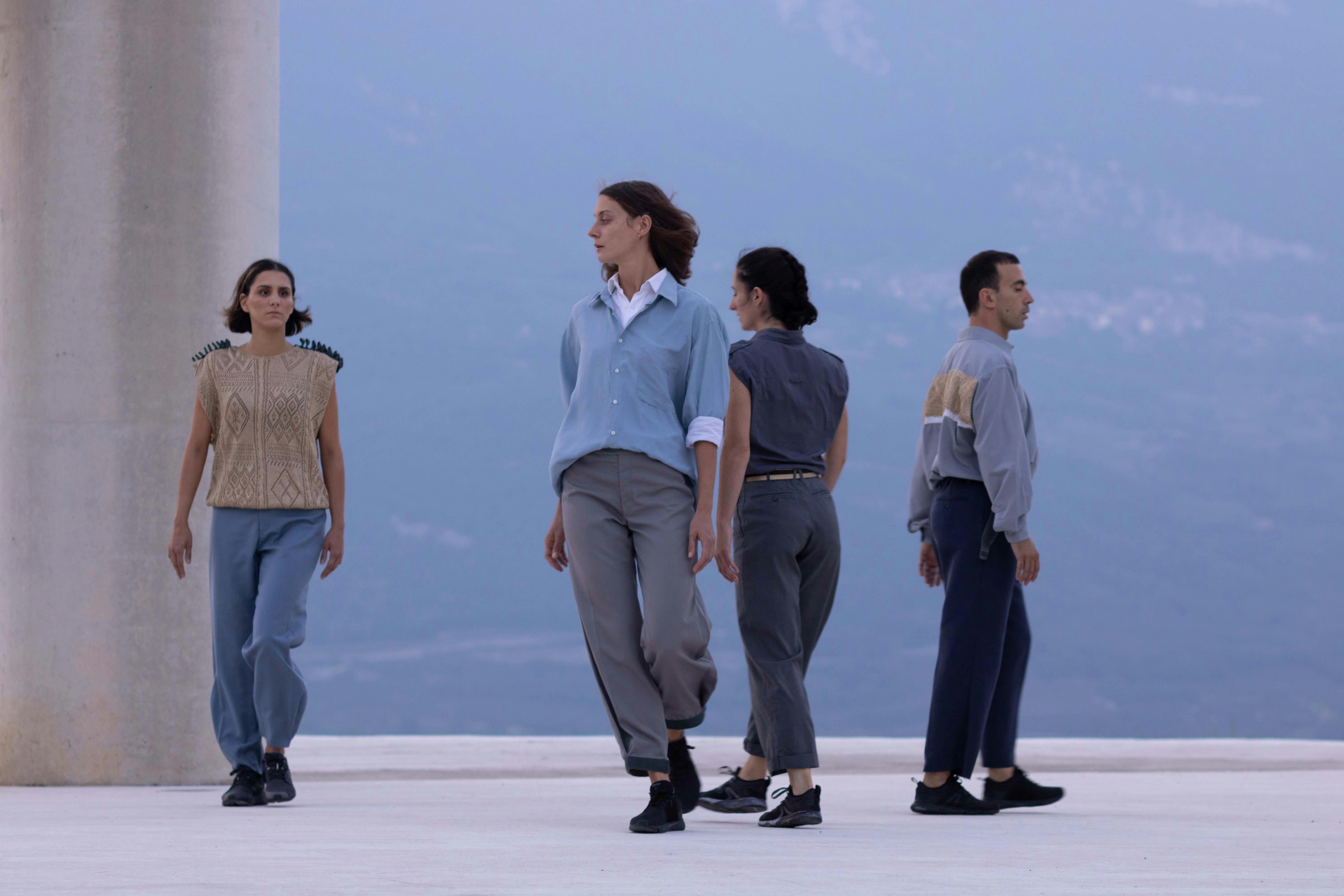 Four dancers walk in the space. Behind them part of a column and in the background the side of a mountain.