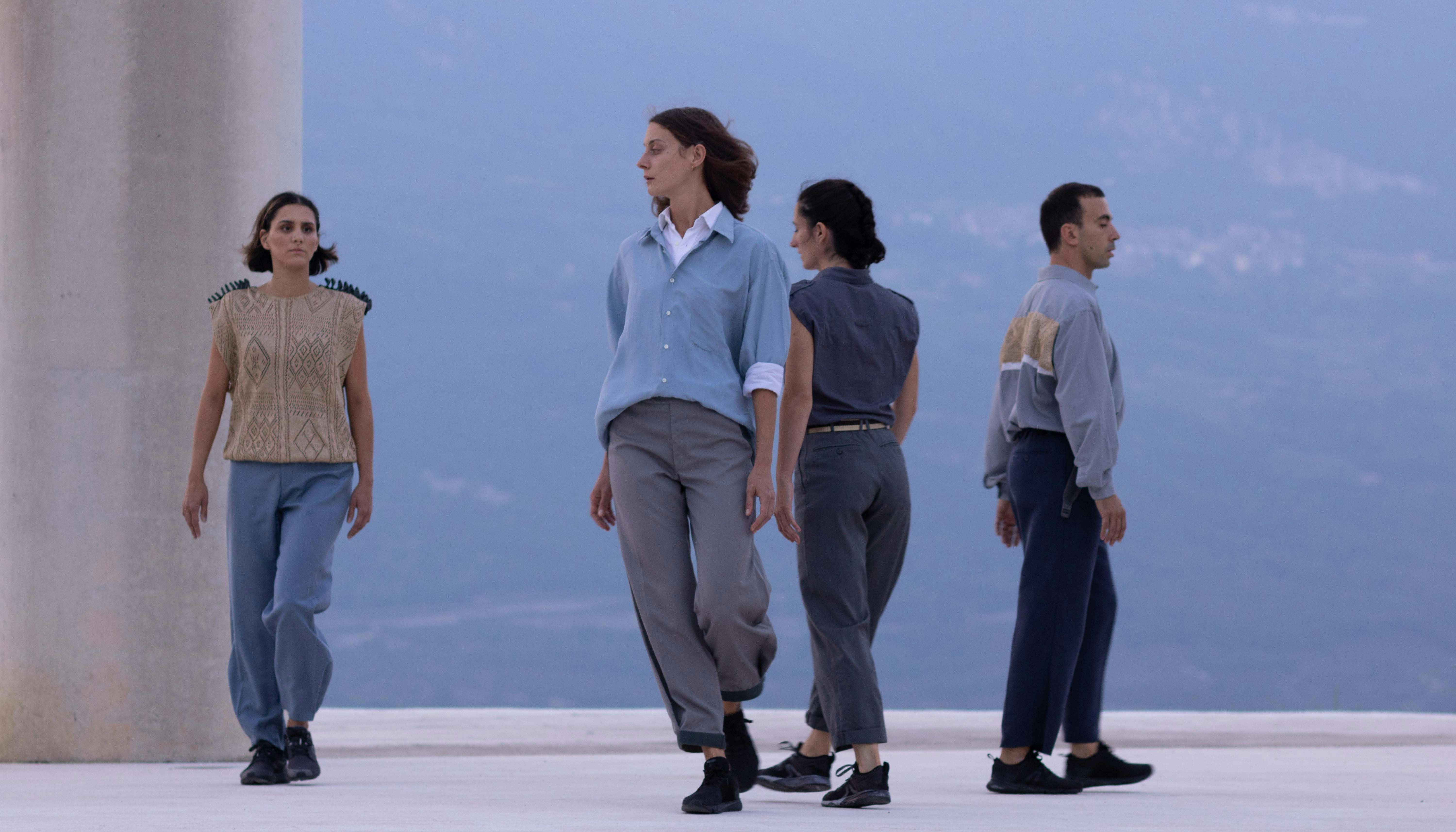 Quattro danzatori e danzatrici camminano nello spazio. Dietro di loro parte di una colonna e sullo sfondo il crinale di una montagna .