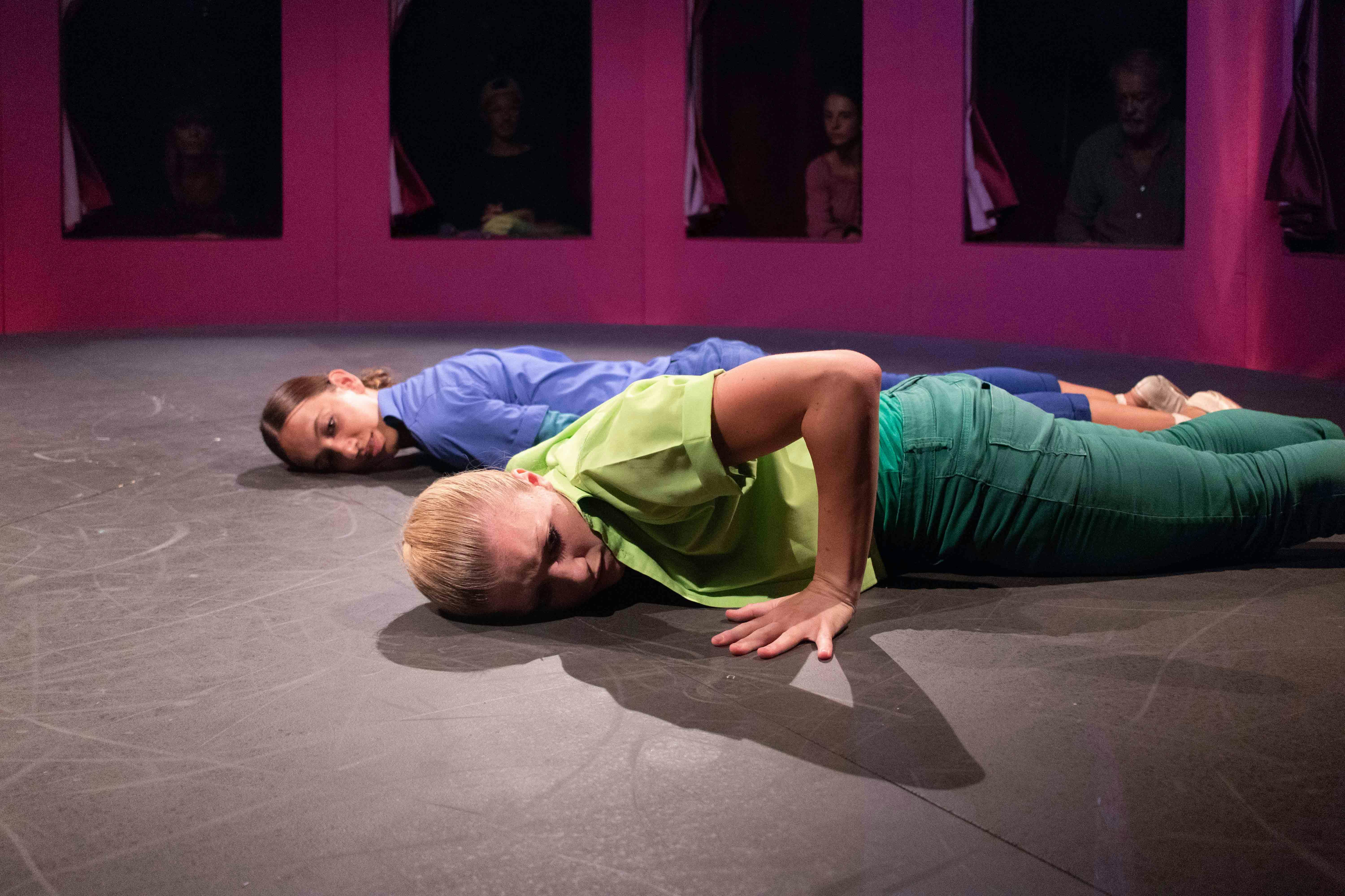 Two dancers are lying prone on the ground, parallel to each other. One is dressed in blue, the other in green. Behind them, through a series of windows, some spectators watch.
