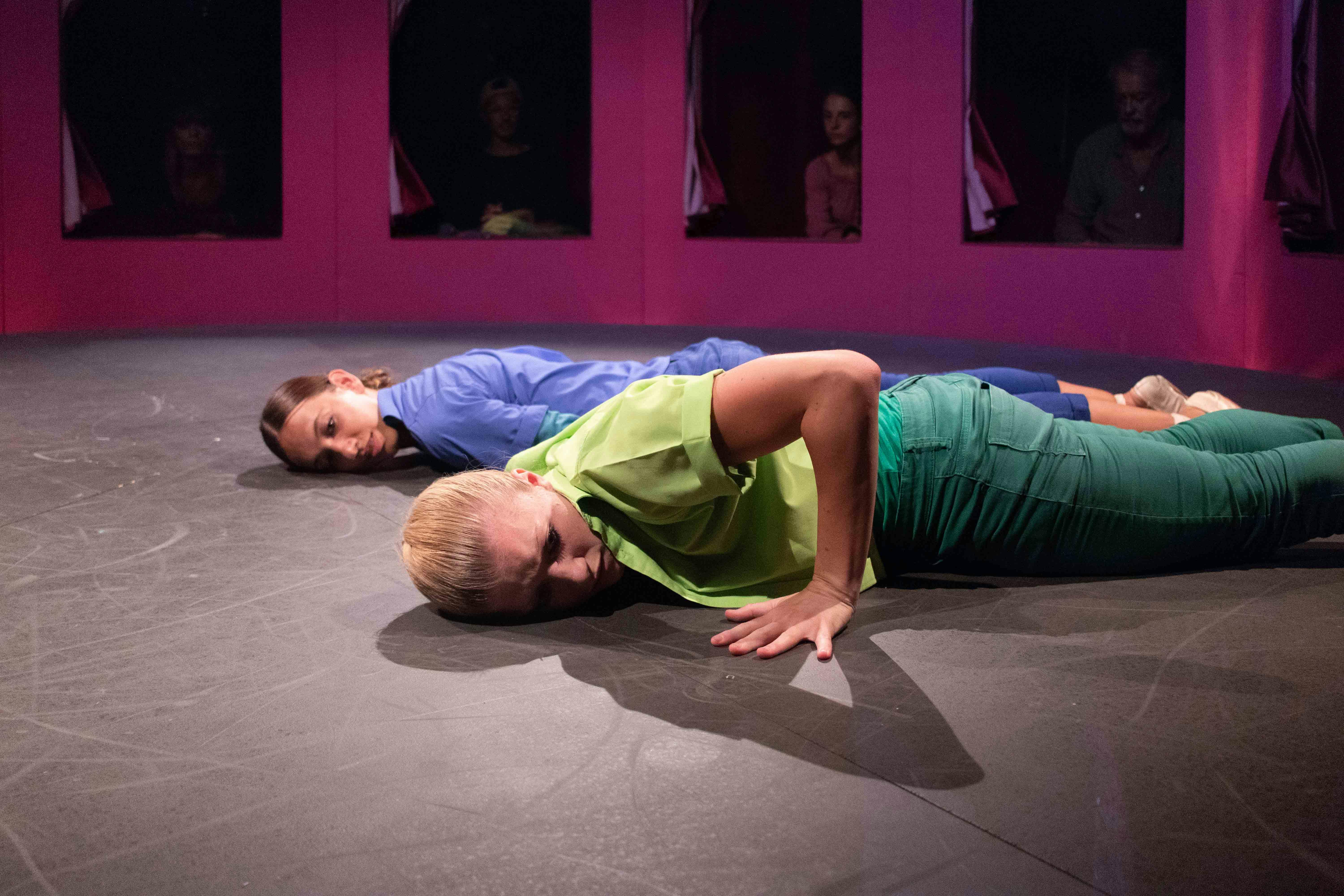 Due danzatrici sono sdraiate a terra prone, parallele tra loro. Una è vestita in blu, l'altra in verde. Dietro di loro, attraverso una serie di finestre, gli spettatori osservano.