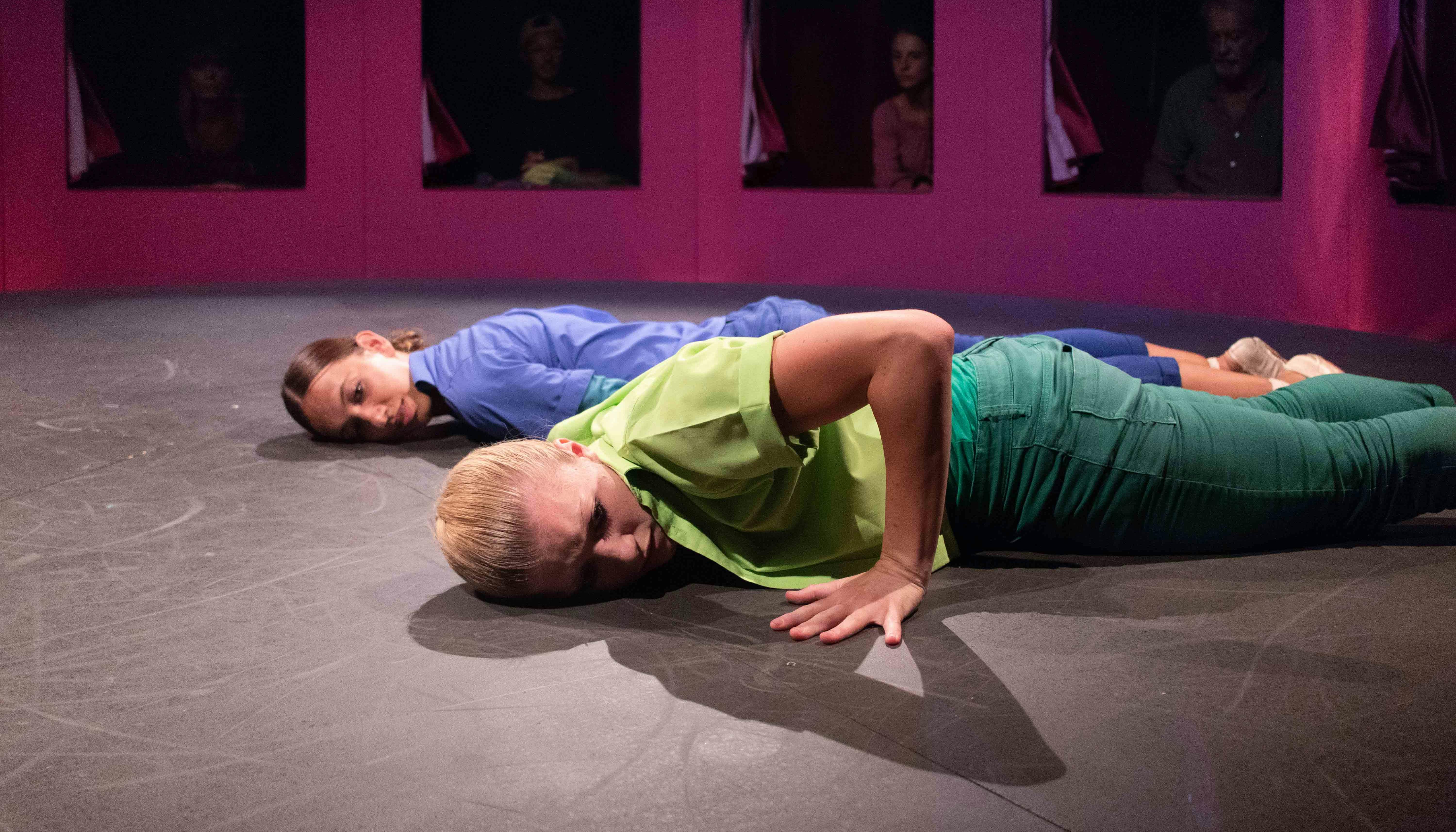 Two dancers are lying prone on the ground, parallel to each other. One is dressed in blue, the other in green. Behind them, through a series of windows, some spectators watch.