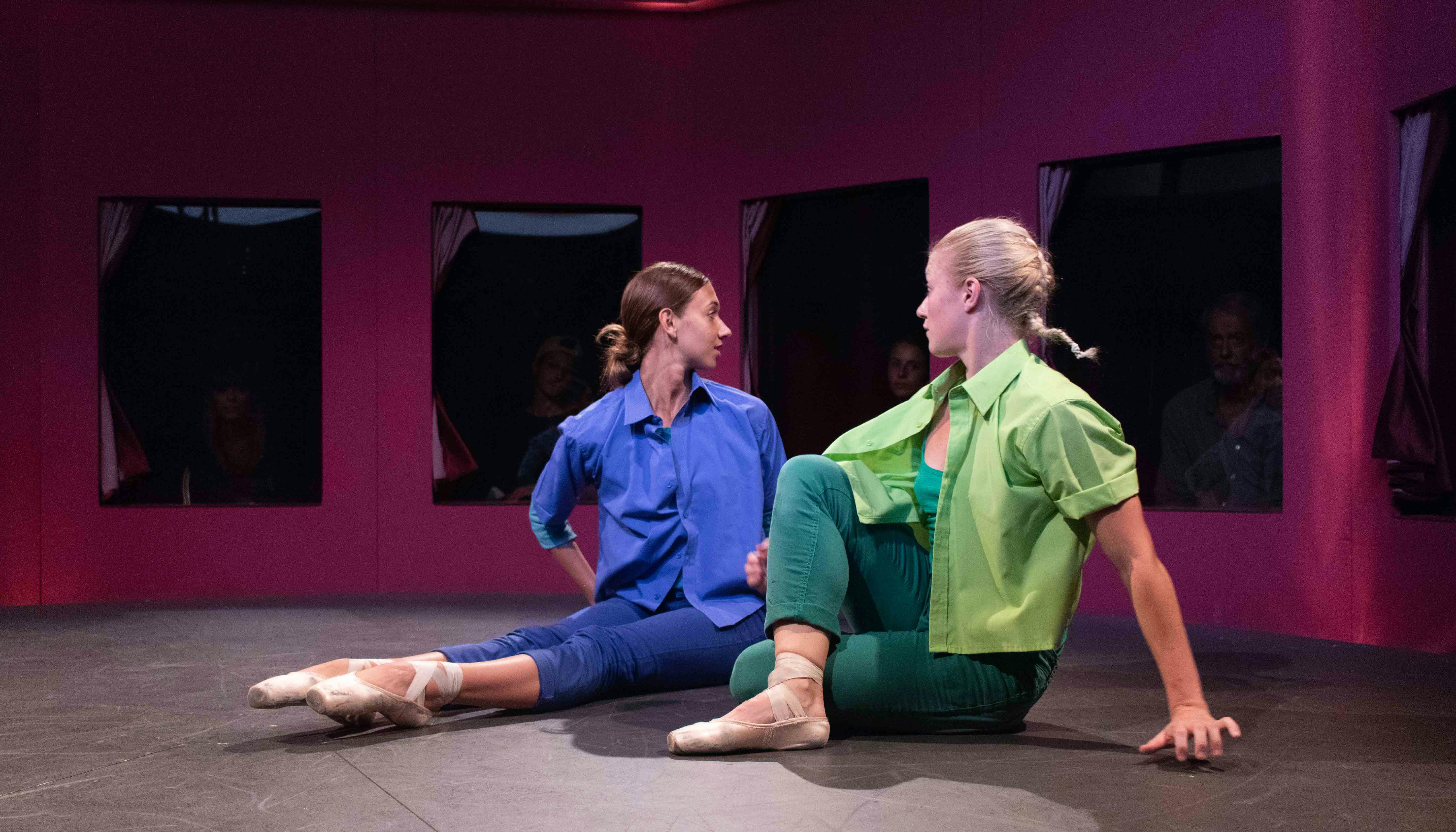 Due danzatrici sono sedute a terra, il busto in torsione all'indietro. Una è vestita in blu, l'altra in verde. Indossano scarpette da punta. Dietro di loro, attraverso una serie di finestre, gli spettatori osservano.