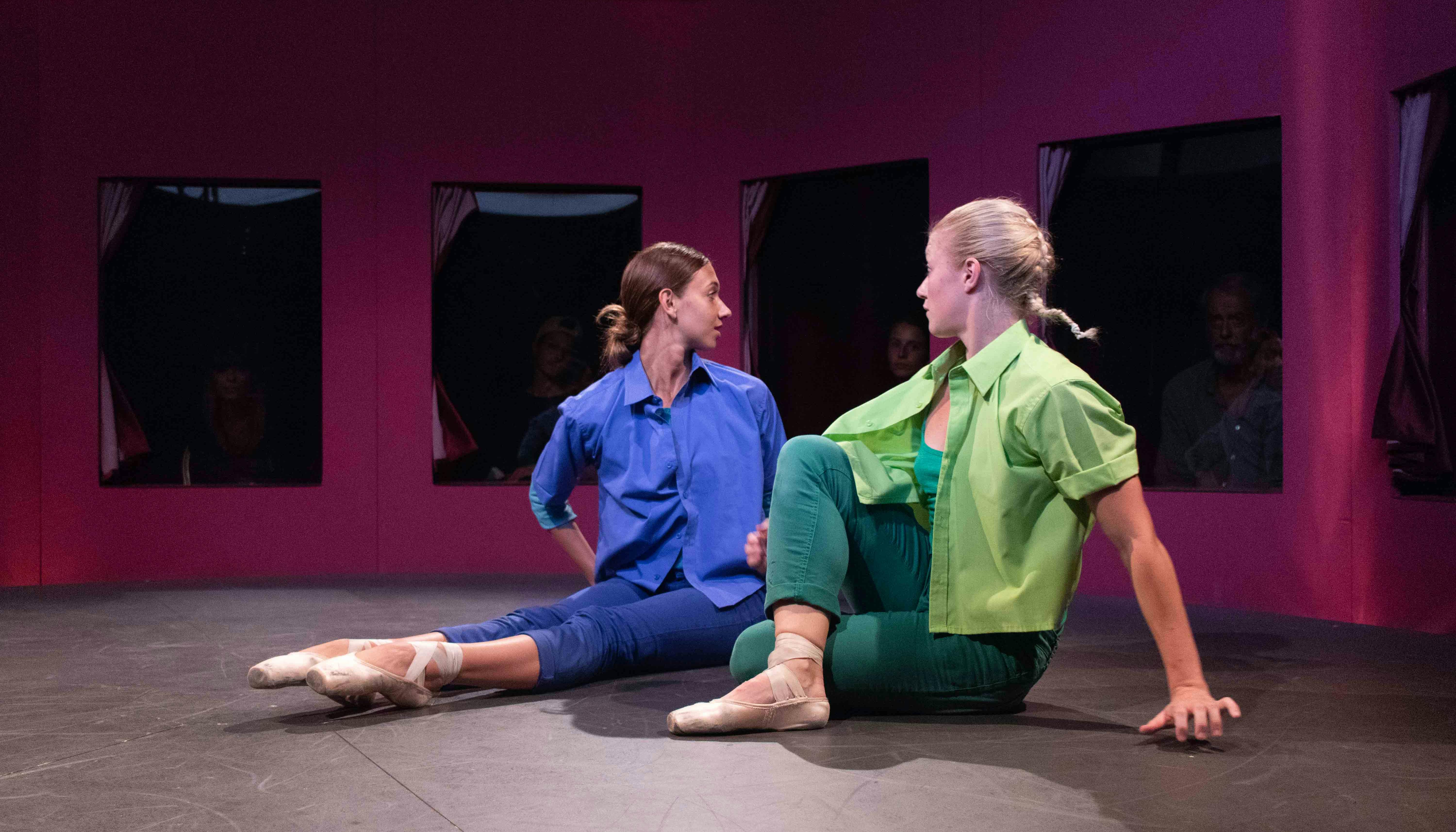 Two dancers are seated on the floor, twisting their torso backwards. One is dressed in blue, the other in green. They are wearing pointe shoes. Behind them, through a series of windows, some spectators watch.