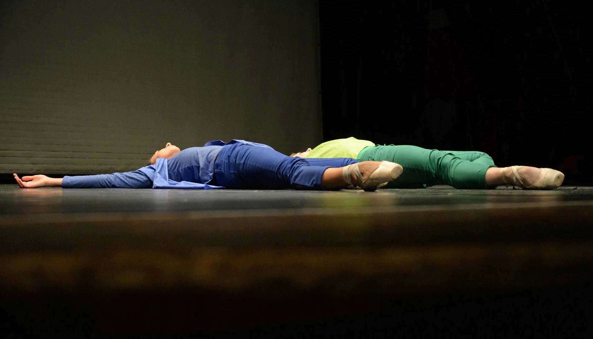 Two dancers are lying on the ground on a stage, parallel to each other. One is dressed in blue, the other one in green. On their feet, facing the audience, they wear pointe shoes.