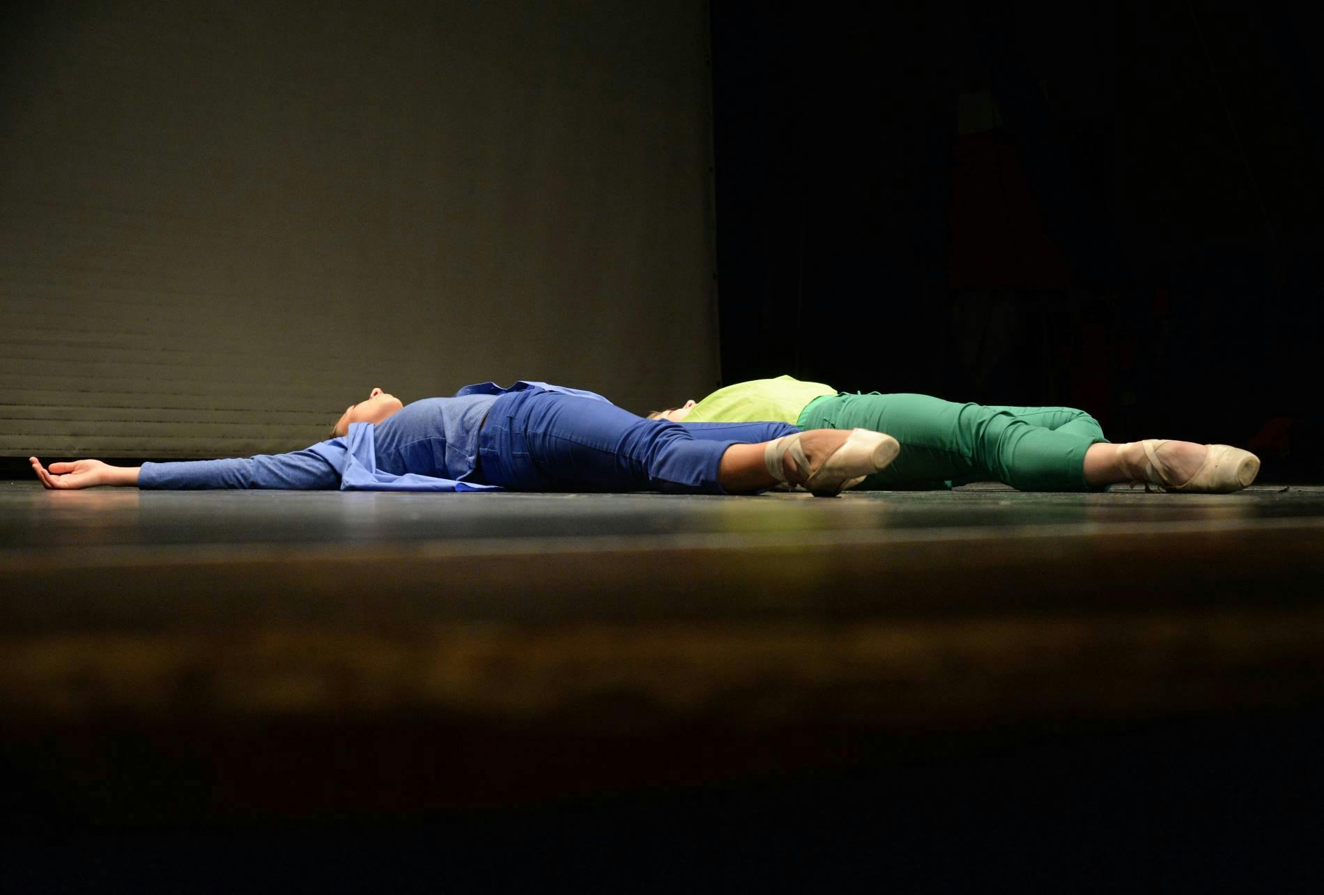 Two dancers are lying on the ground on a stage, parallel to each other. One is dressed in blue, the other one in green. On their feet, facing the audience, they wear pointe shoes.