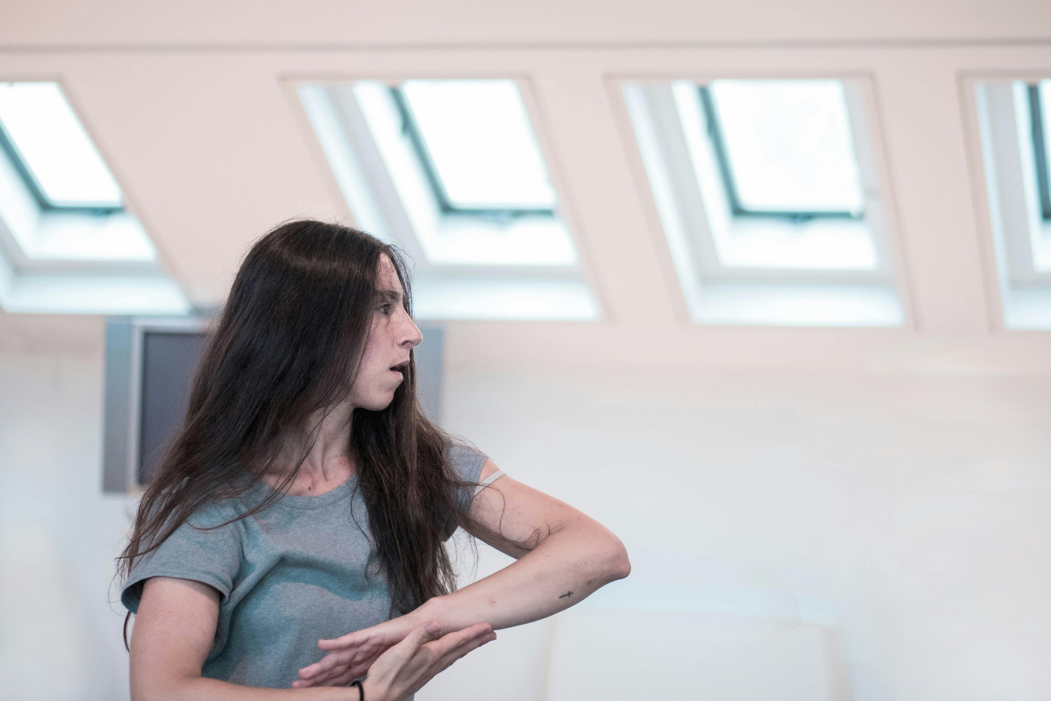 Dancer Sara Sguotti, wearing a gray T-shirt during a rehearsal. Her elbows are bent and her hands overlap in front of her torso.