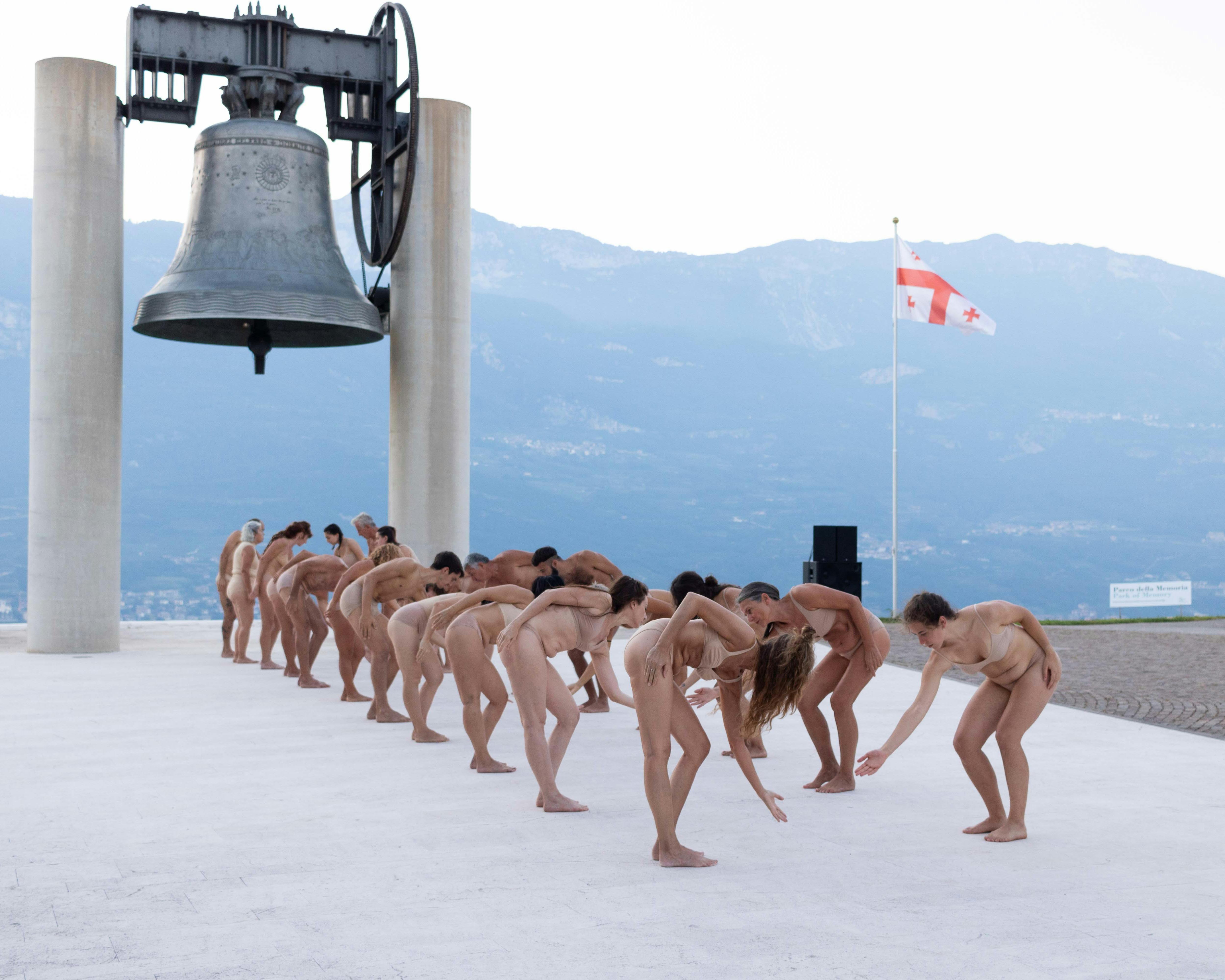 A group of nude dancers, with different physicalities, are standing on two parallel lines, facing each other.  They are moving, leaning forward with their torso. Behind them we see the monument Campana dei Caduti and some mountains in the background.