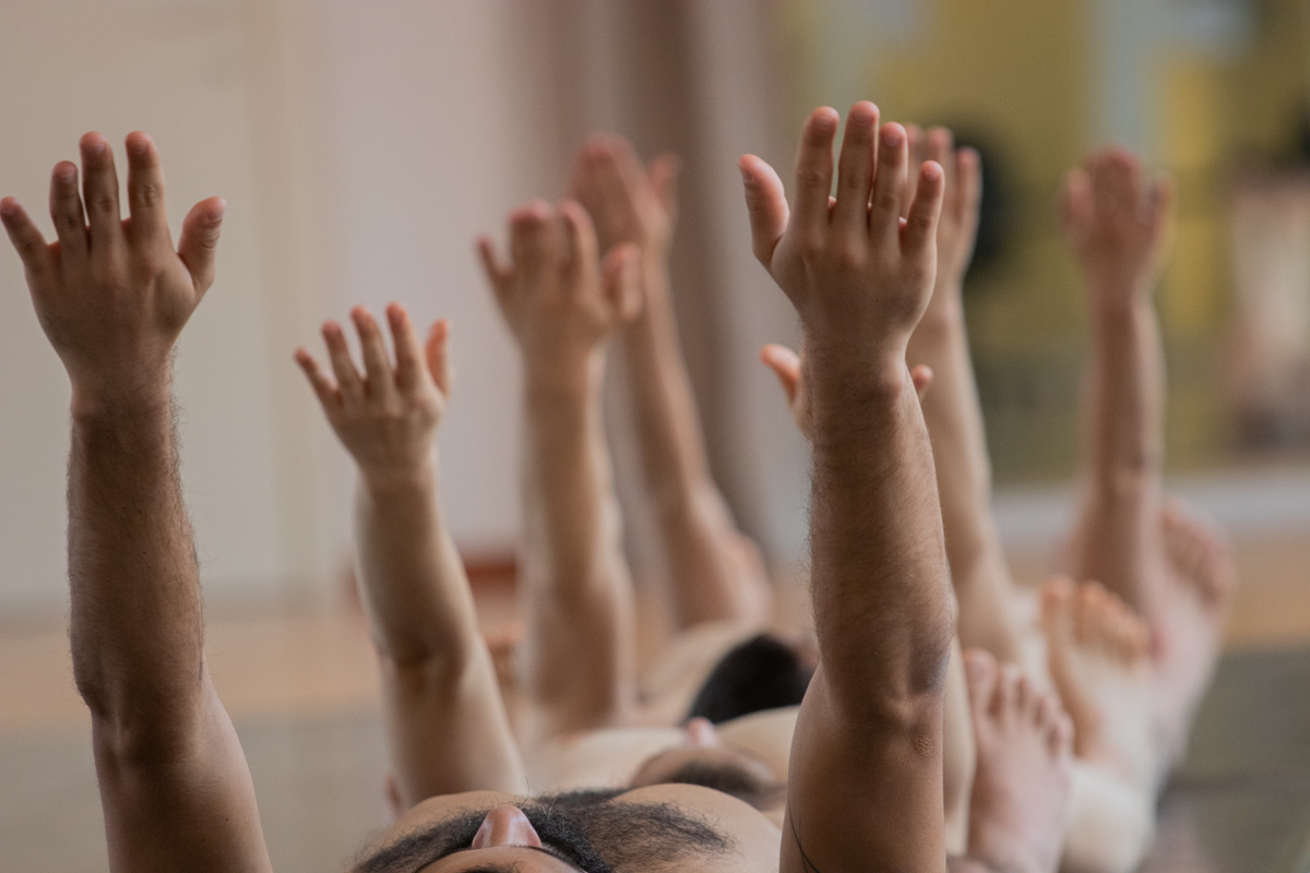 Four dancers are lying on their backs on the floor, their arms stretched upwards.