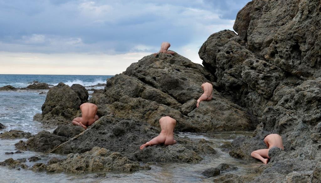 Una scogliera rocciosa sul mare. Cinque corpi umani nudi sono seduti di schiena tra le roccie. Il cielo è nuvoloso.