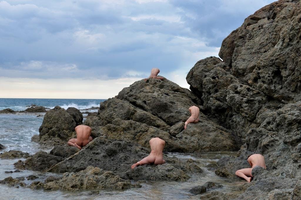 A cliff on the sea. Five naked human bodies are sitting among the rocks with their backs to the camera.  The sky is cloudy.