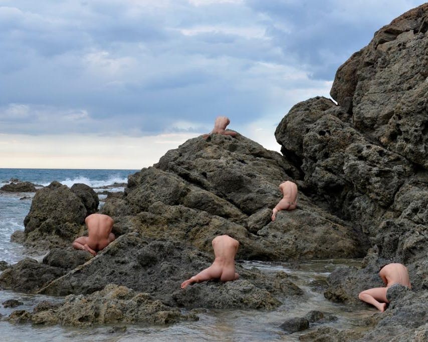 A cliff on the sea. Five naked human bodies are sitting among the rocks with their backs to the camera.  The sky is cloudy.