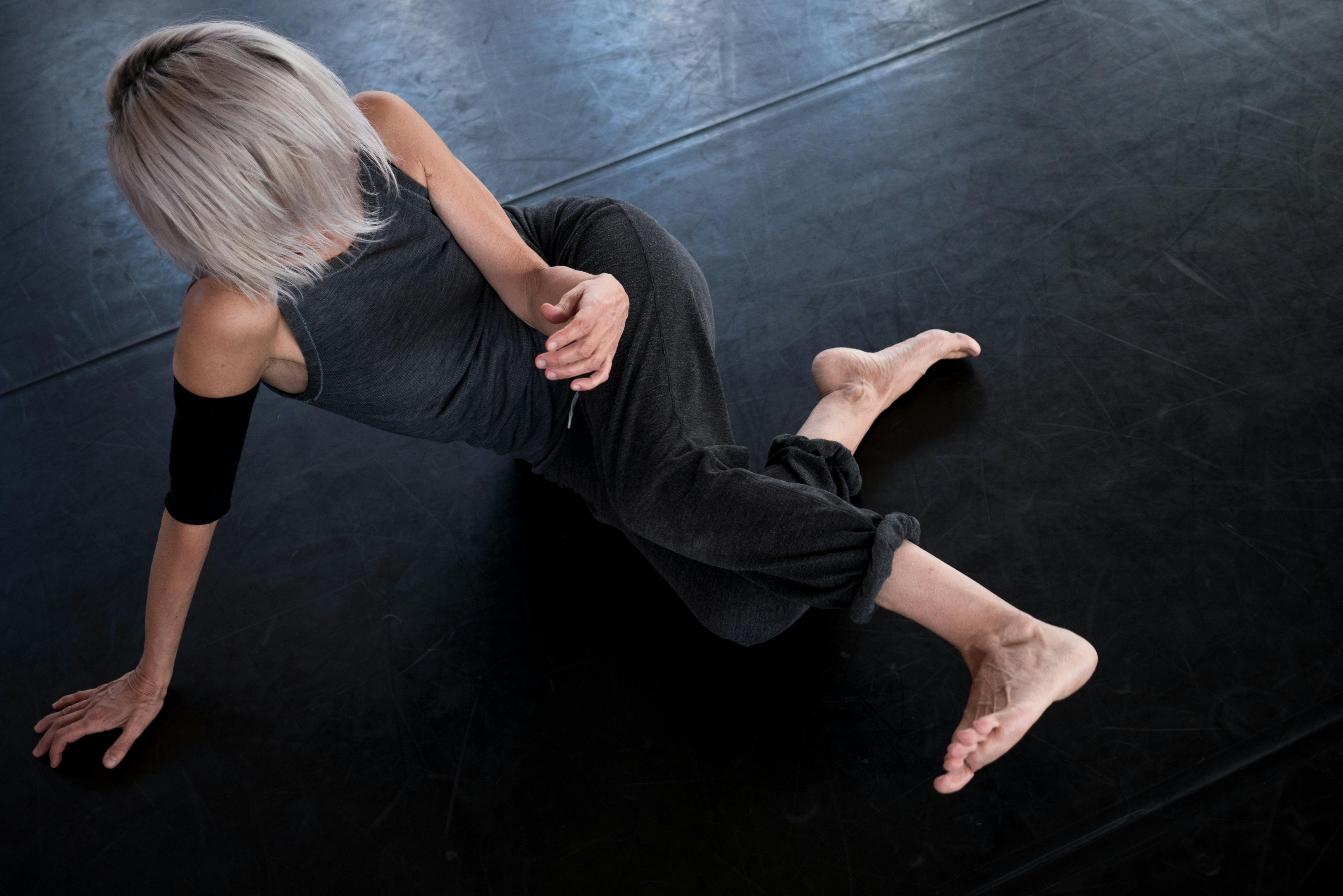 Paola Bianchi, dressed in black, in torsion on the floor. She lifts part of her body, supporting her weight on her right arm. Her shoulder-length, very light hair covers her face.